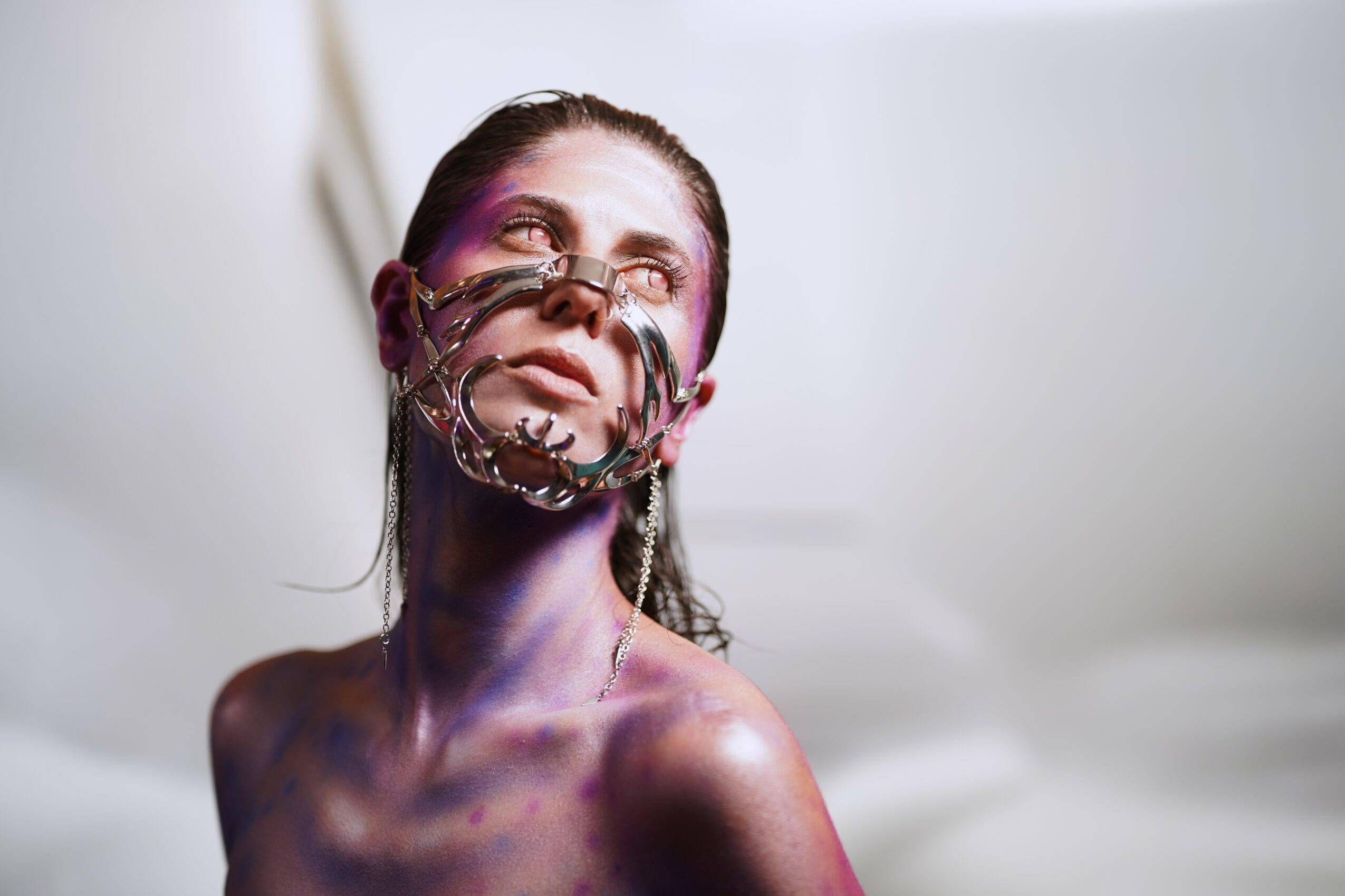 Woman with makeup and a decorative face mask, looking upward, with a neutral background.