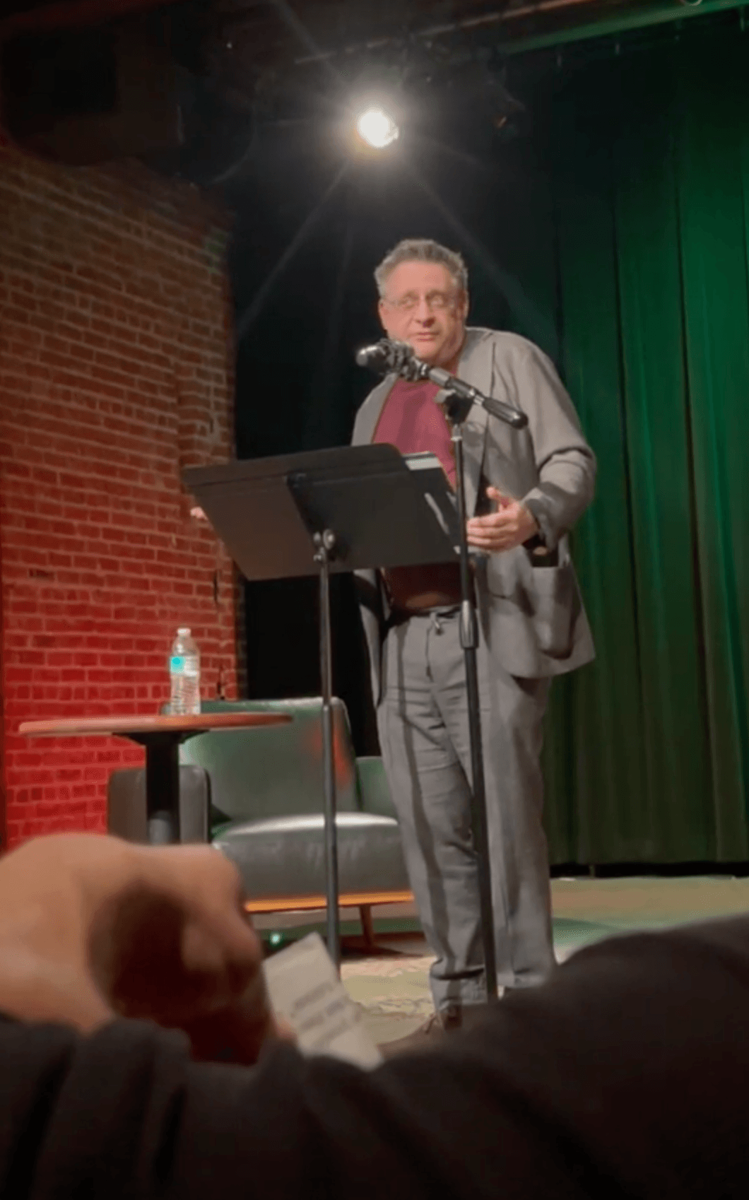 Man speaking at a podium on stage with green curtain background, brick wall, and a water bottle on a small table.