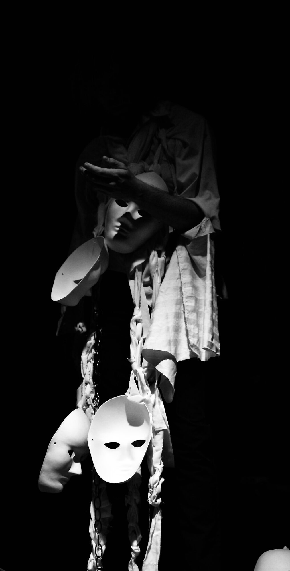 Person holding a mask and hanging masks, with a dark background, in black and white.