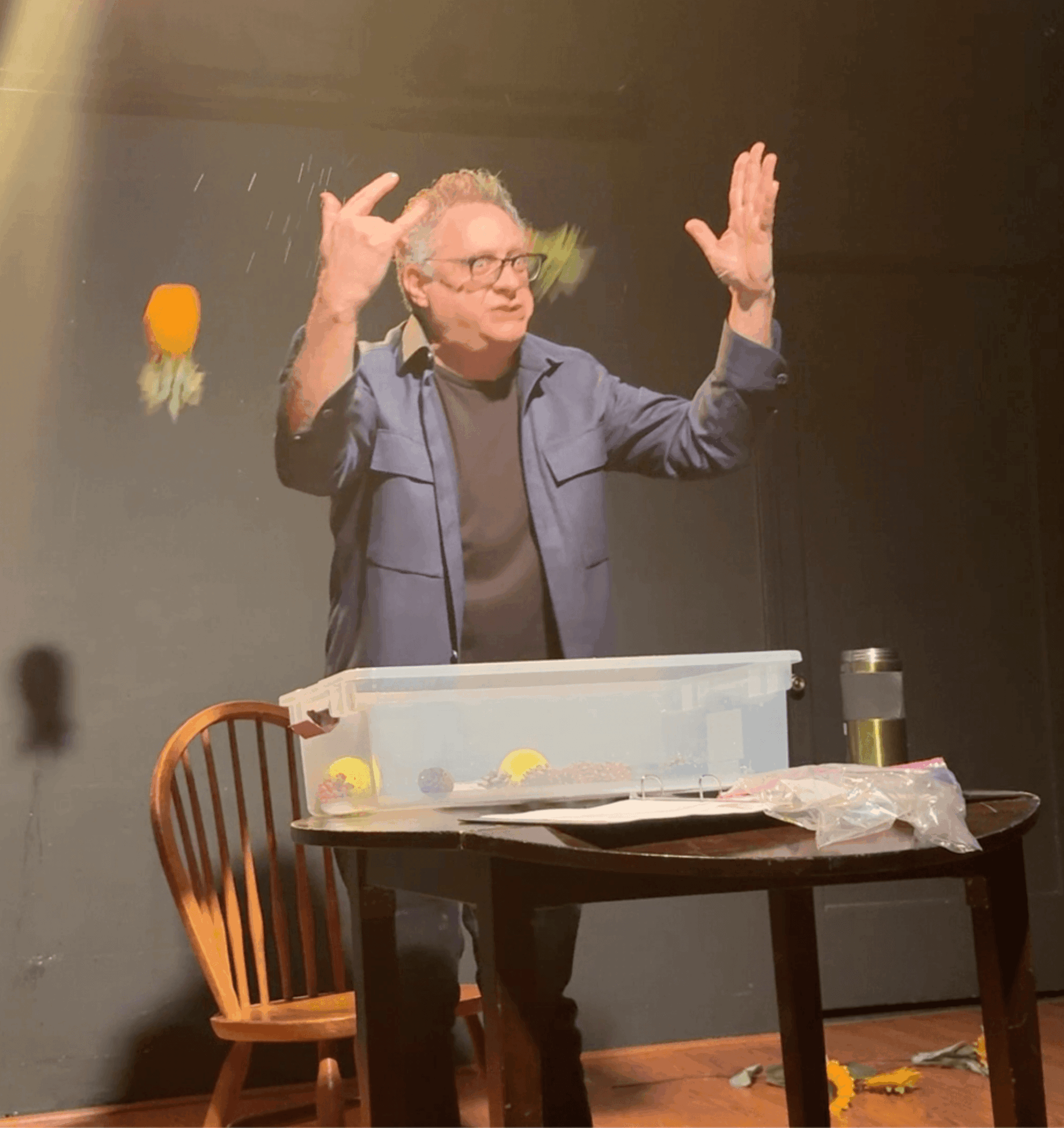 Person with glasses and gray hair, wearing a blue jacket, standing behind a table with objects, raising hands, in a room.