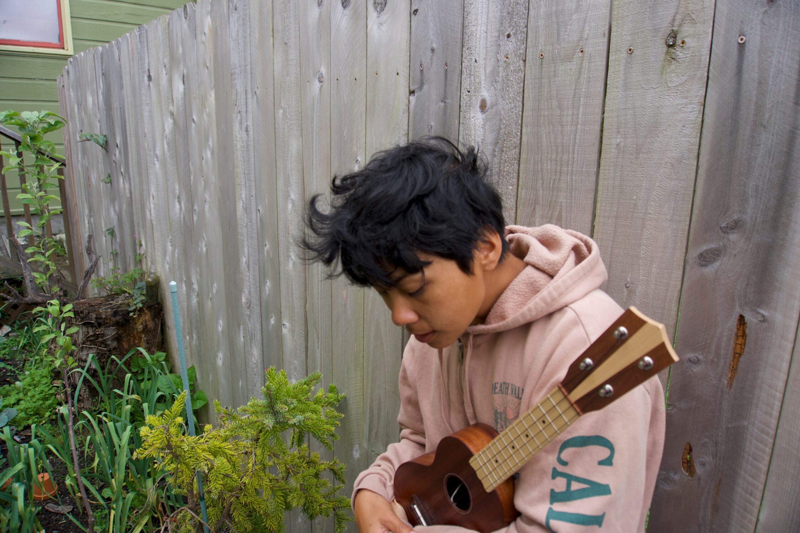 Young person with dark curly hair wearing a pink hoodie, holding a small guitar outdoors near a wooden fence.