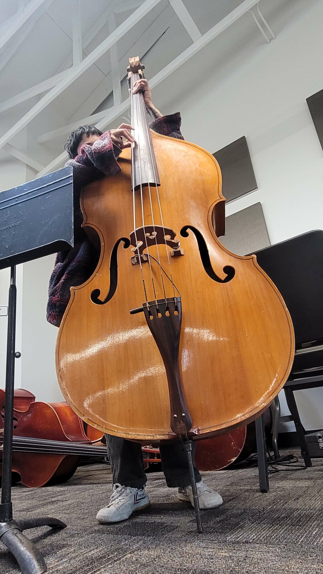 Person playing a large wooden double bass, with a music stand nearby, in an indoor setting.