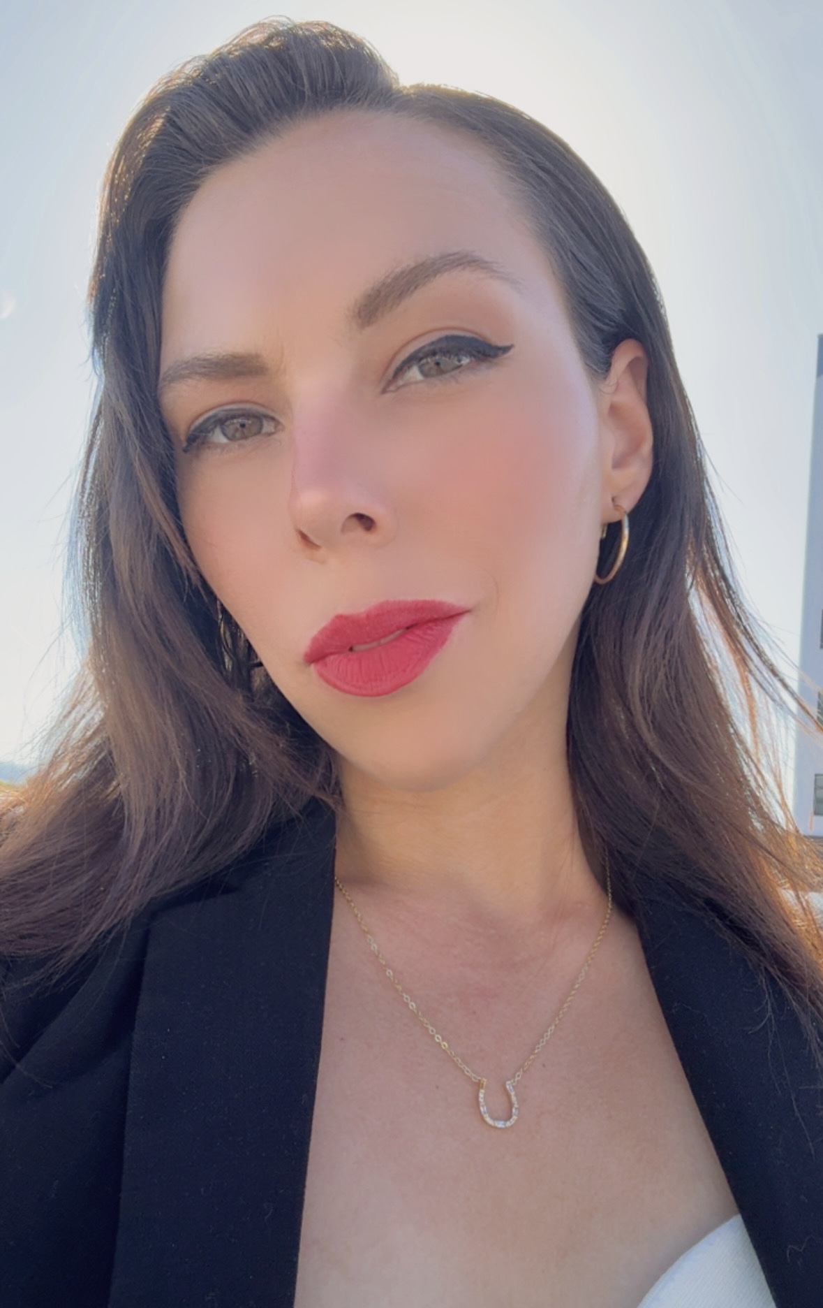 Close-up of a woman with makeup, red lipstick, and long hair, wearing a black blazer and a necklace.