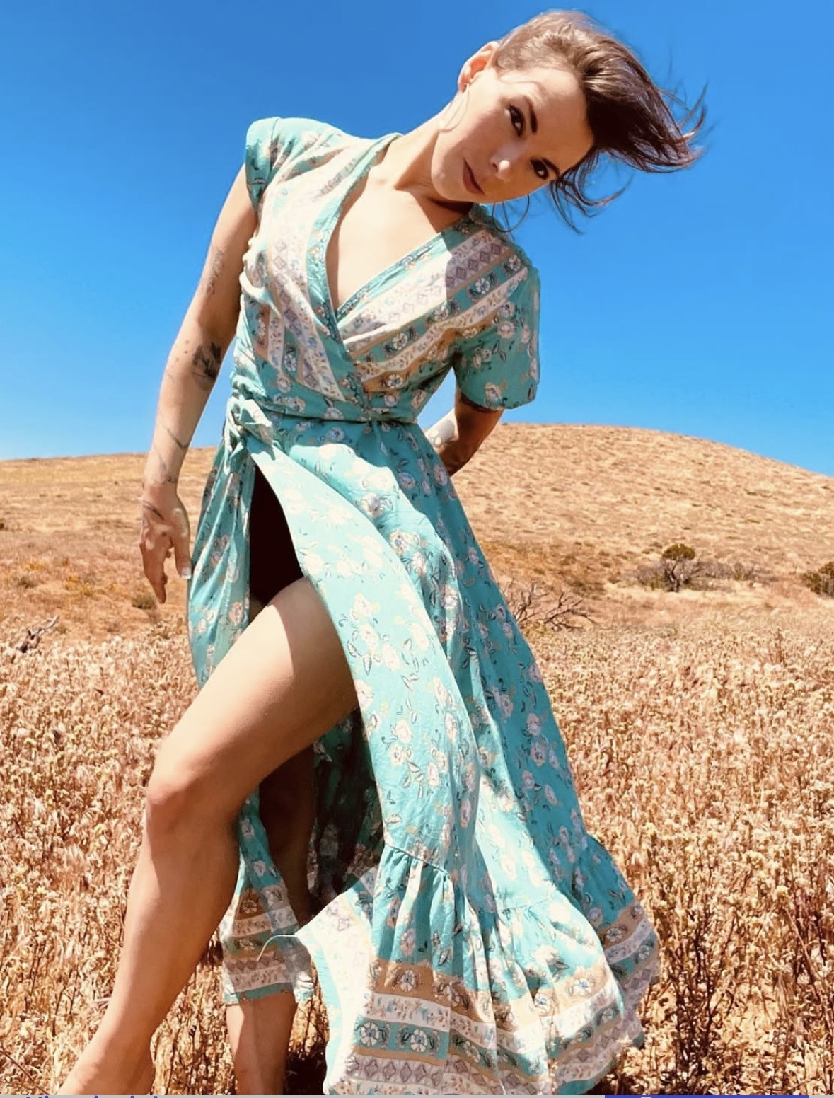 Woman in a flowing dress standing in a dry field under a clear blue sky, leaning forward with her hair blowing.