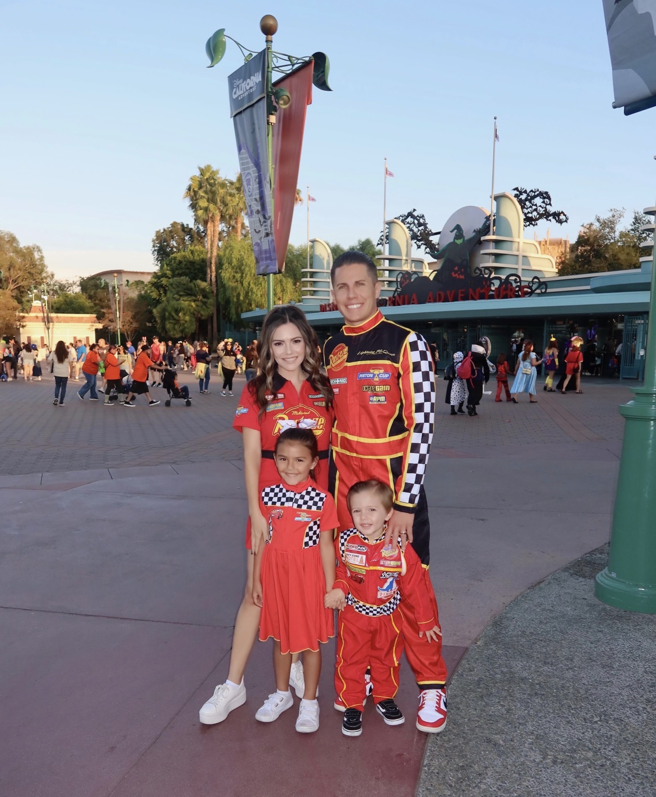 A family of three in racing costumes at an amusement park, with a crowd and park buildings in the background.