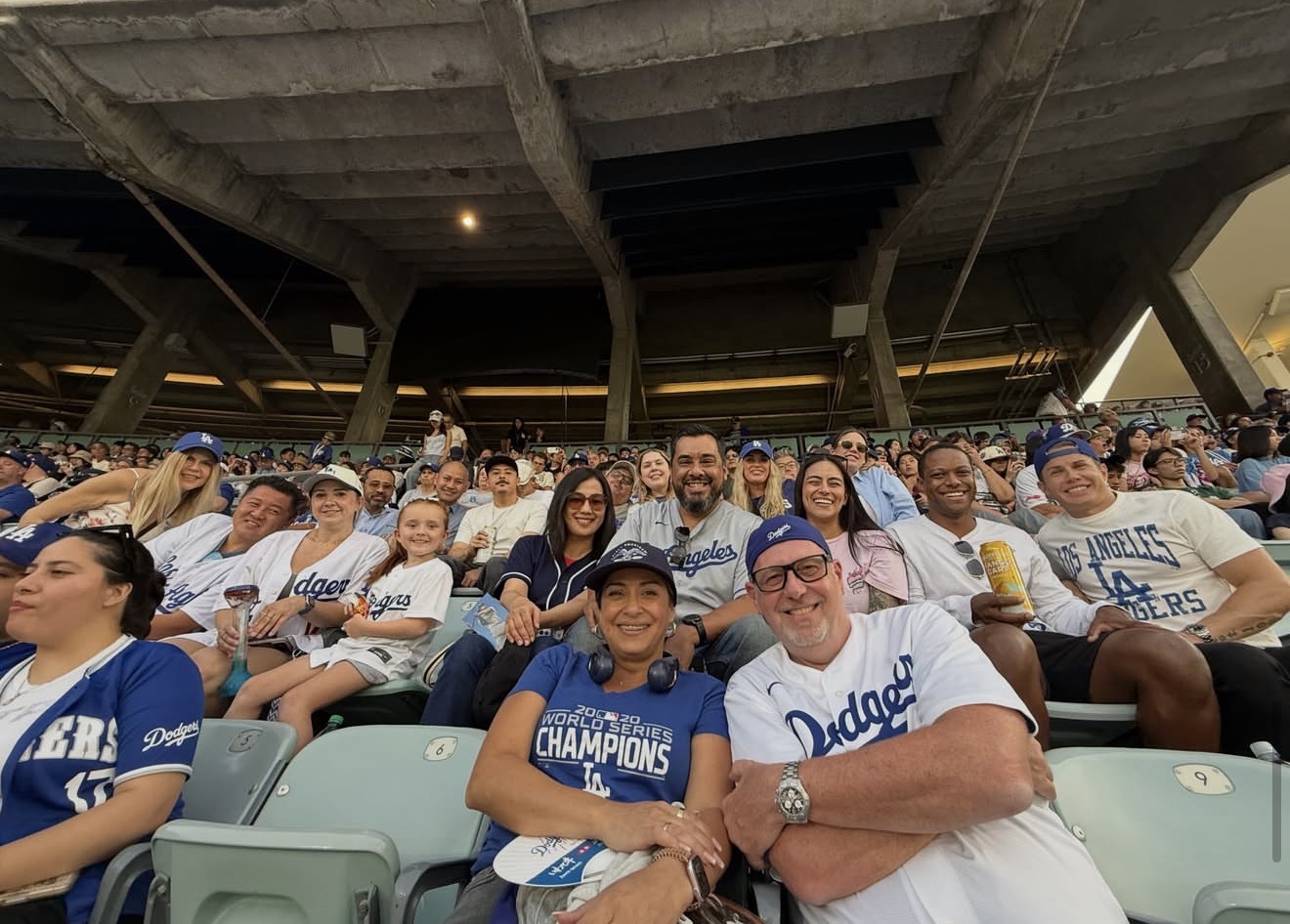 Crowd of people sitting in stadium seats, some wearing Dodgers shirts, under a large stadium roof.
