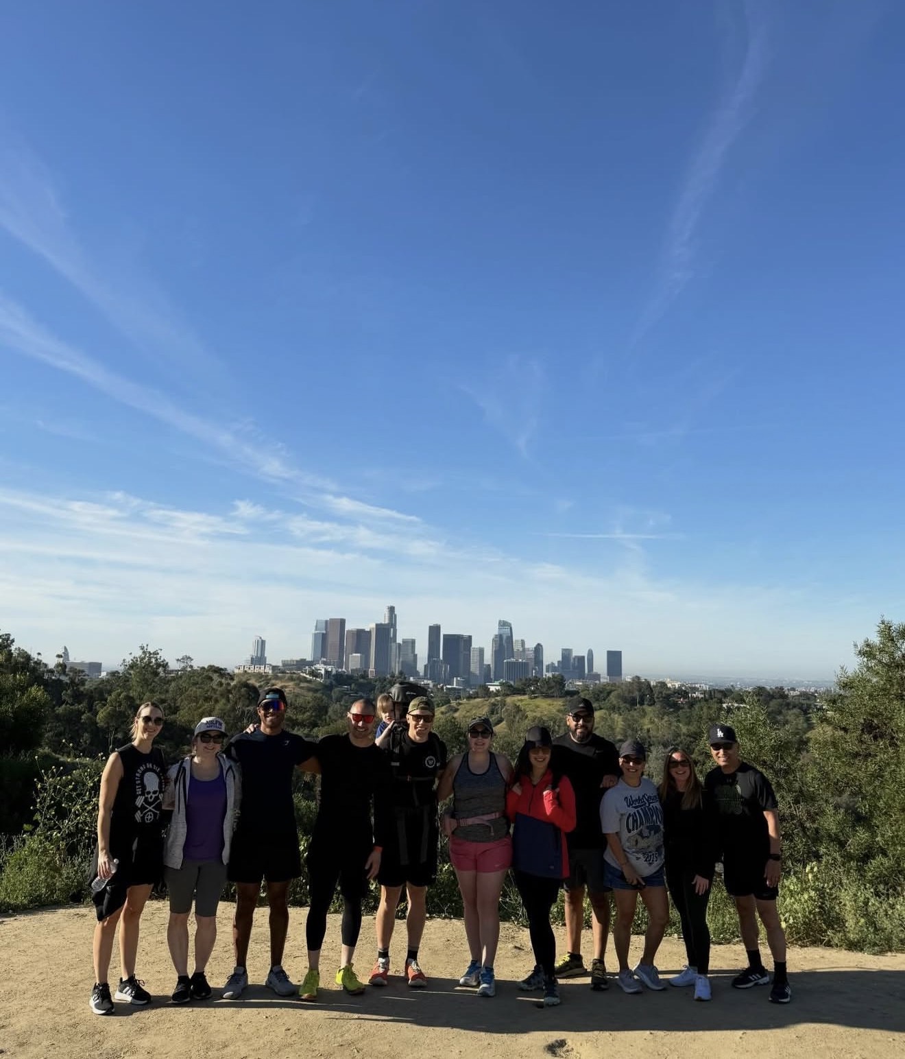 Group of ten people standing outdoors with city skyline in background, clear blue sky, some trees, casual clothing, smiling.