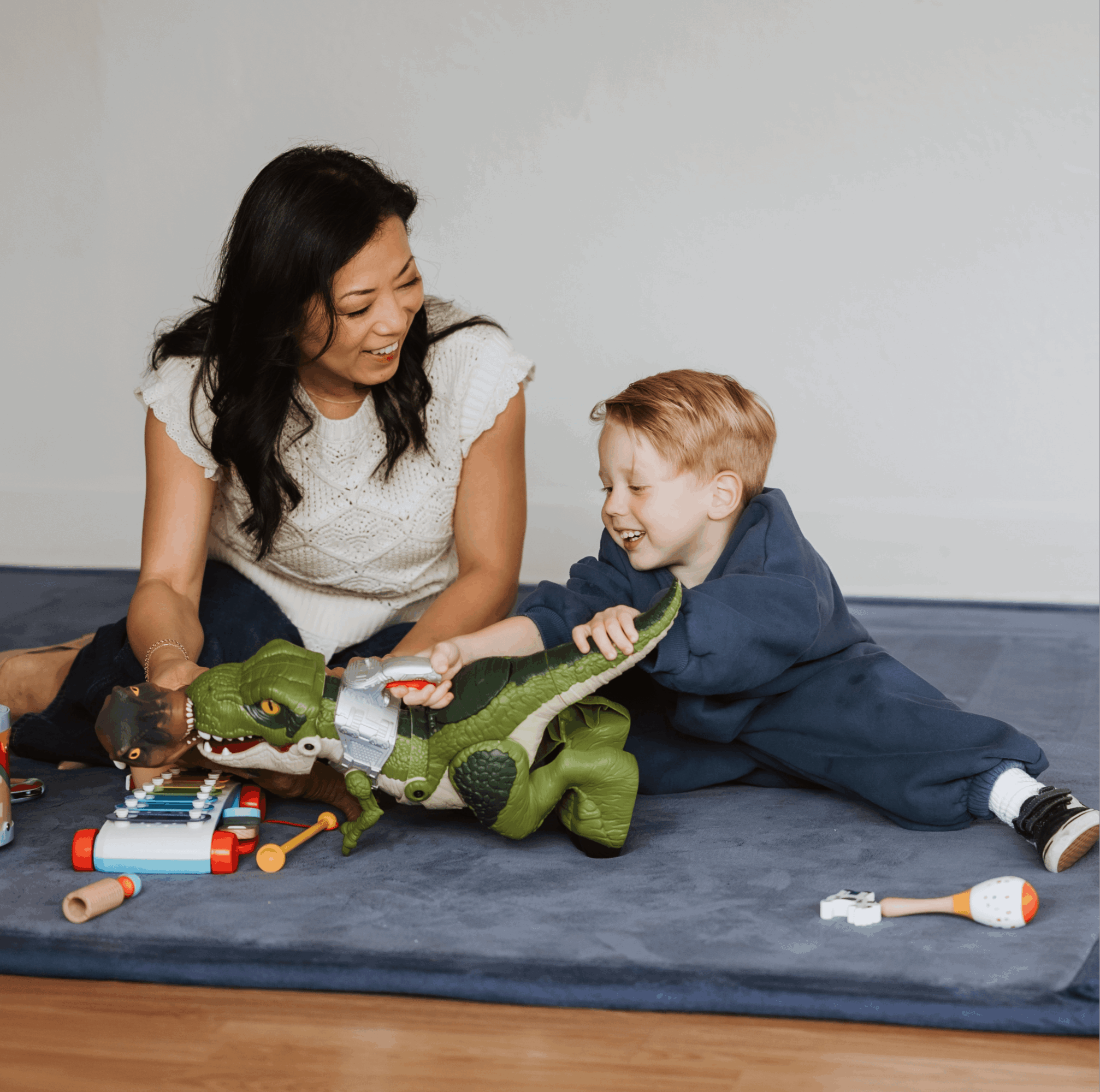 Woman and child playing with a green toy crocodile on a blue mat, smiling and engaging with toys nearby.