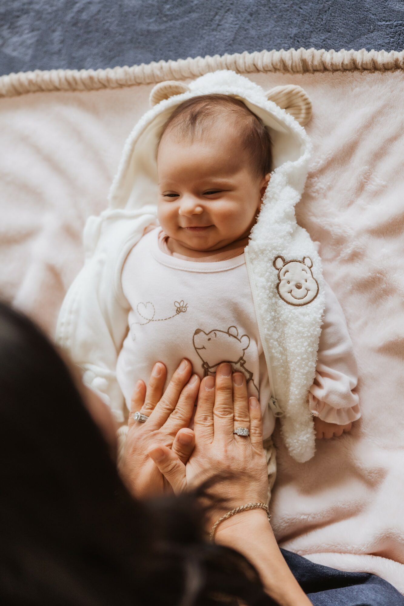 Baby lying on a blanket, wearing a bear hat and a onesie with animal illustrations, being touched by an adult's hands.