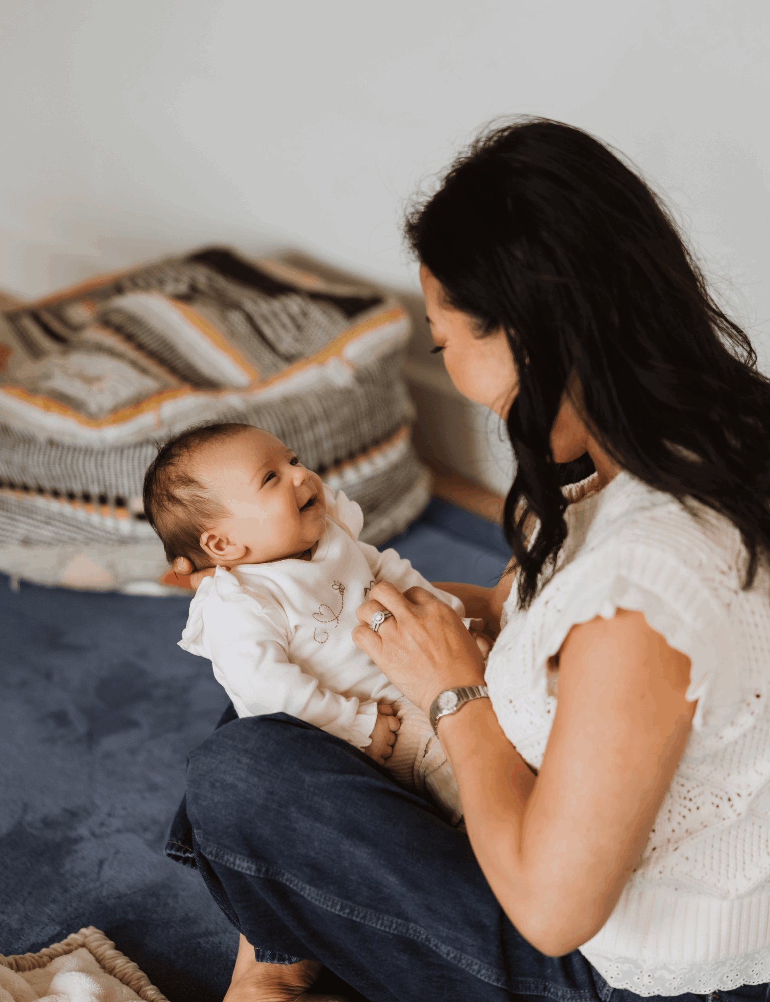 Woman holding a baby and looking at each other indoors, with a bed and pillows in the background.