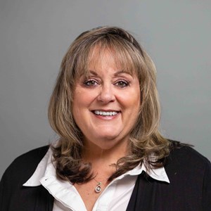 Smiling woman with shoulder-length blonde hair, wearing a white shirt and black blazer, against a gray background.