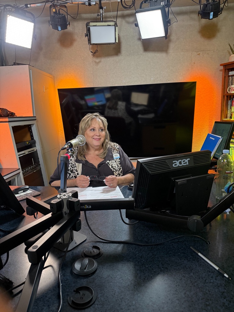 Woman sitting at a desk with computer monitors, microphone, and studio lights overhead in a recording studio.