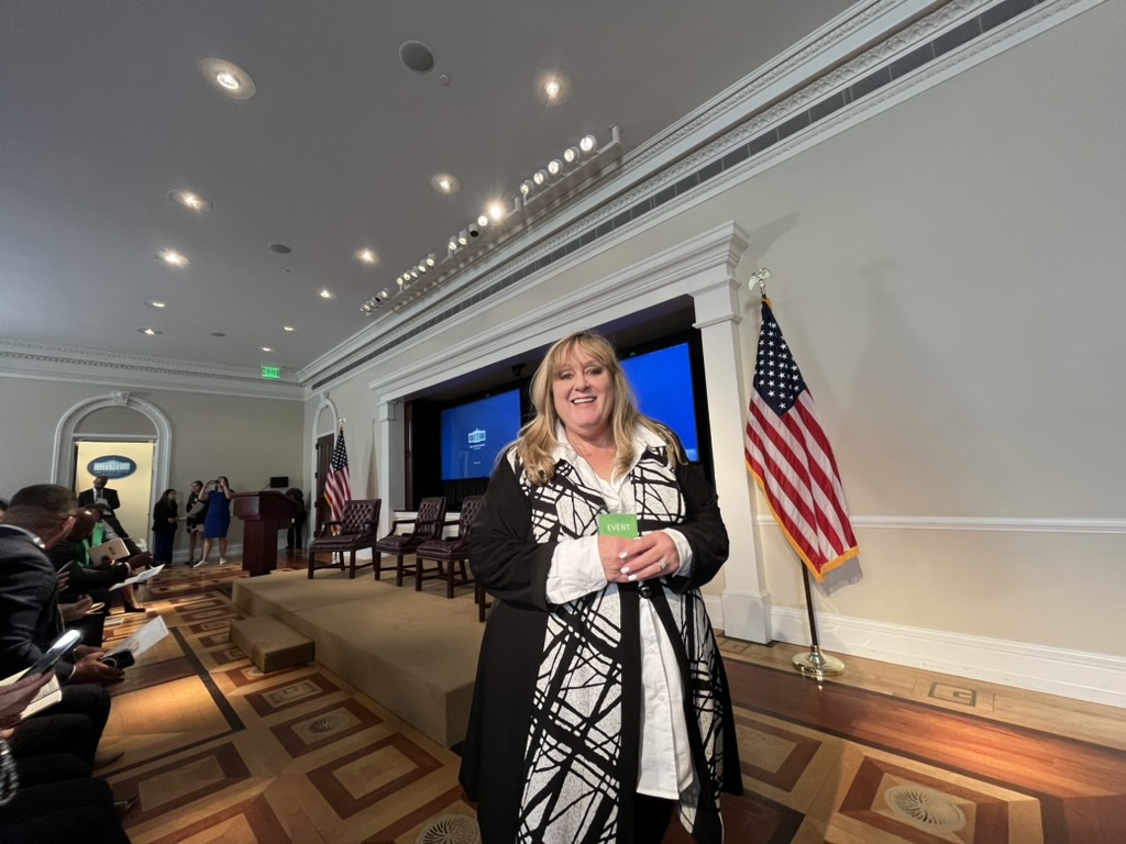 Smiling woman holding a green card, standing in front of a stage with American flag and large screen, in a formal room.