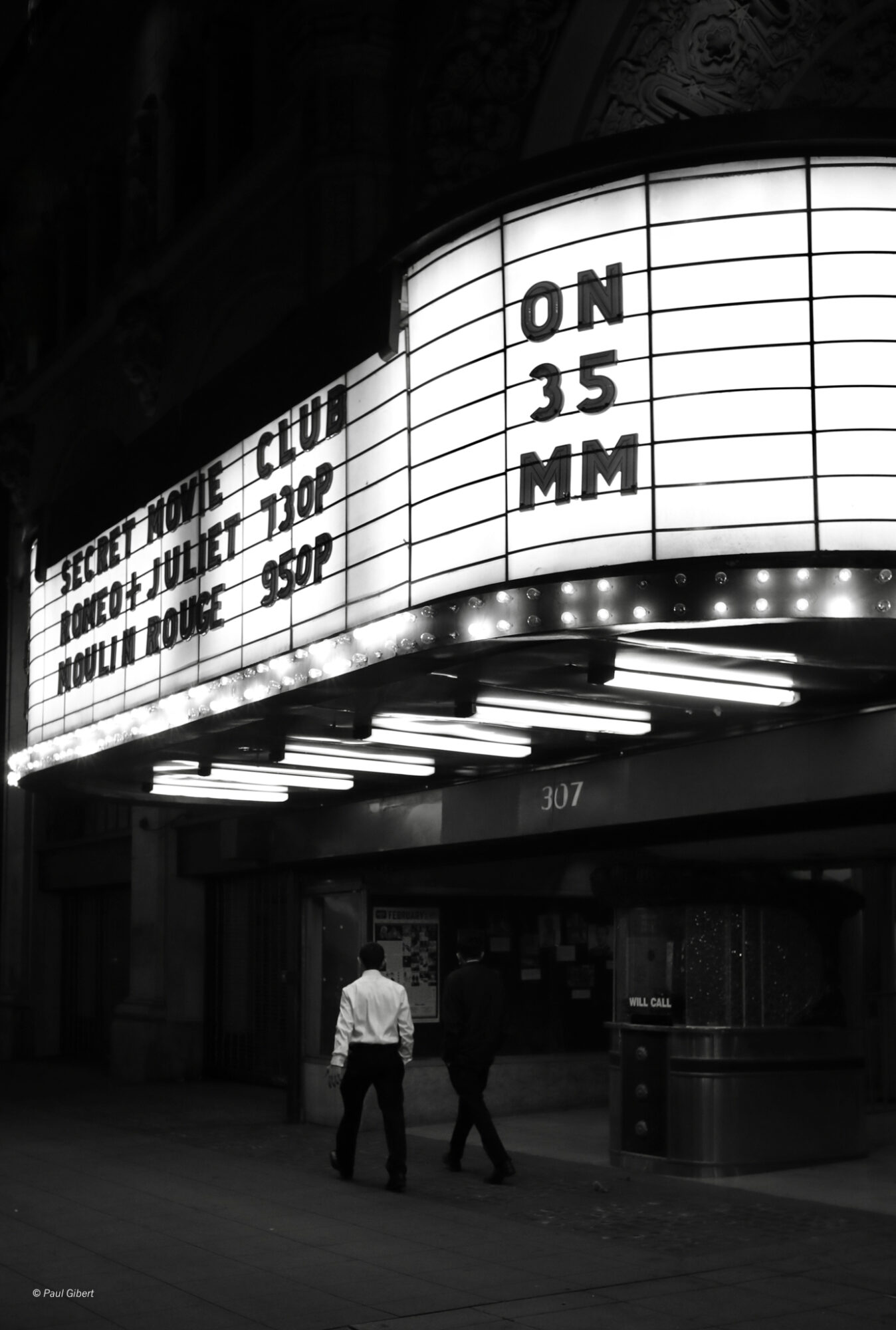 People walking past a lit movie theater marquee displaying movie titles and showtimes at night.