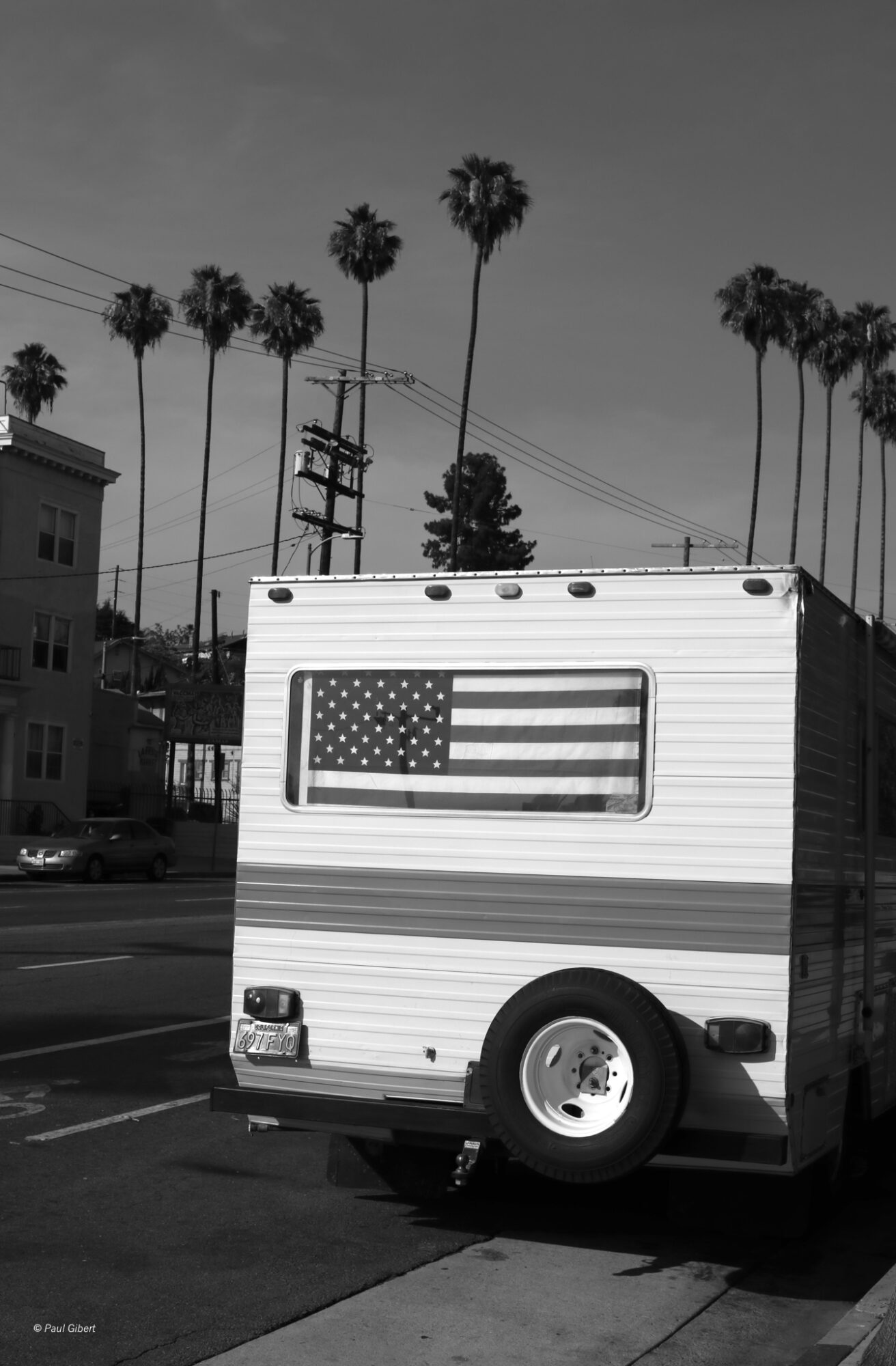 A black and white image of a trailer with an American flag design on its side, parked on a street with palm trees in the background.