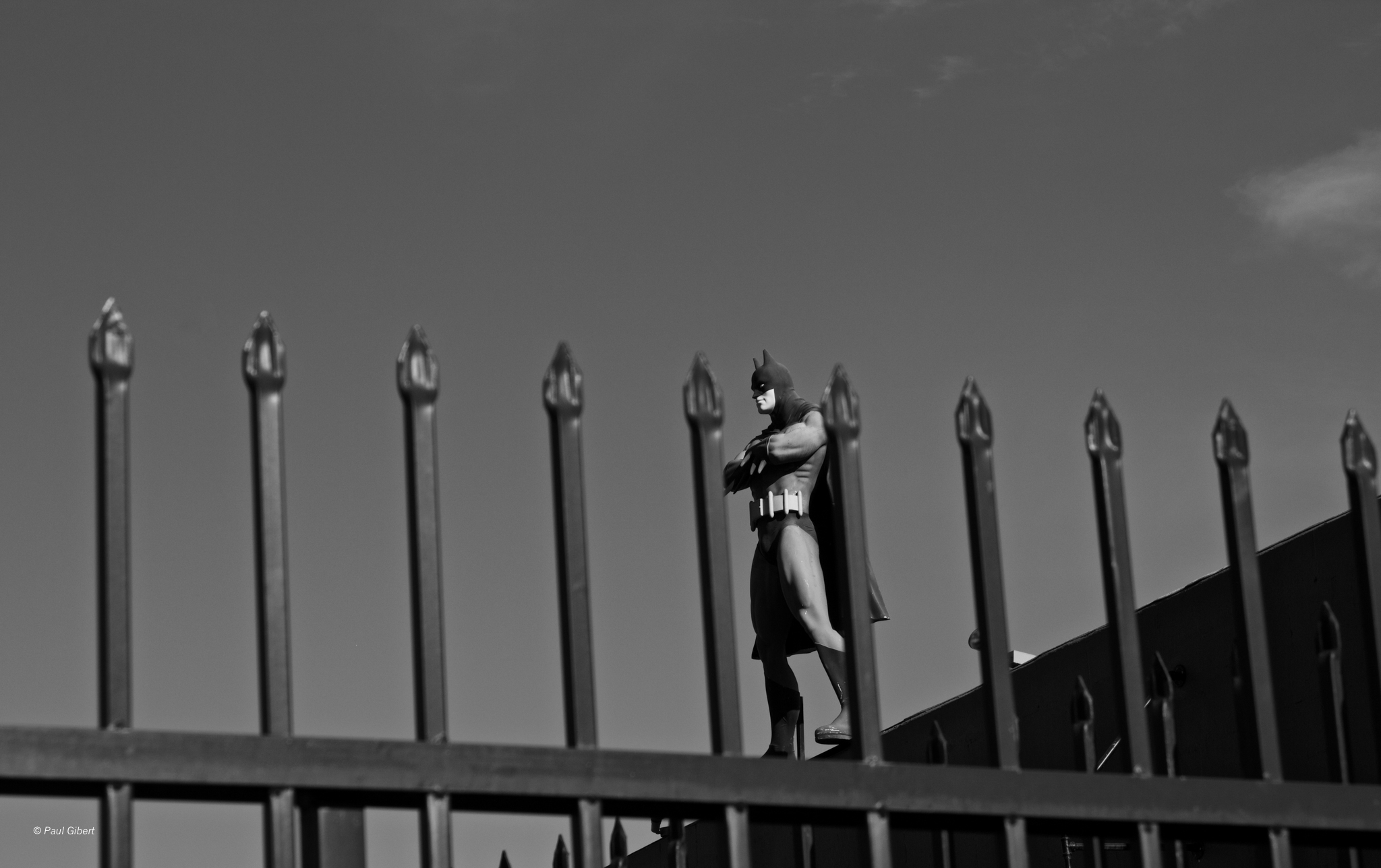 Person standing on a fence, holding a phone, with a building in the background, in black and white.