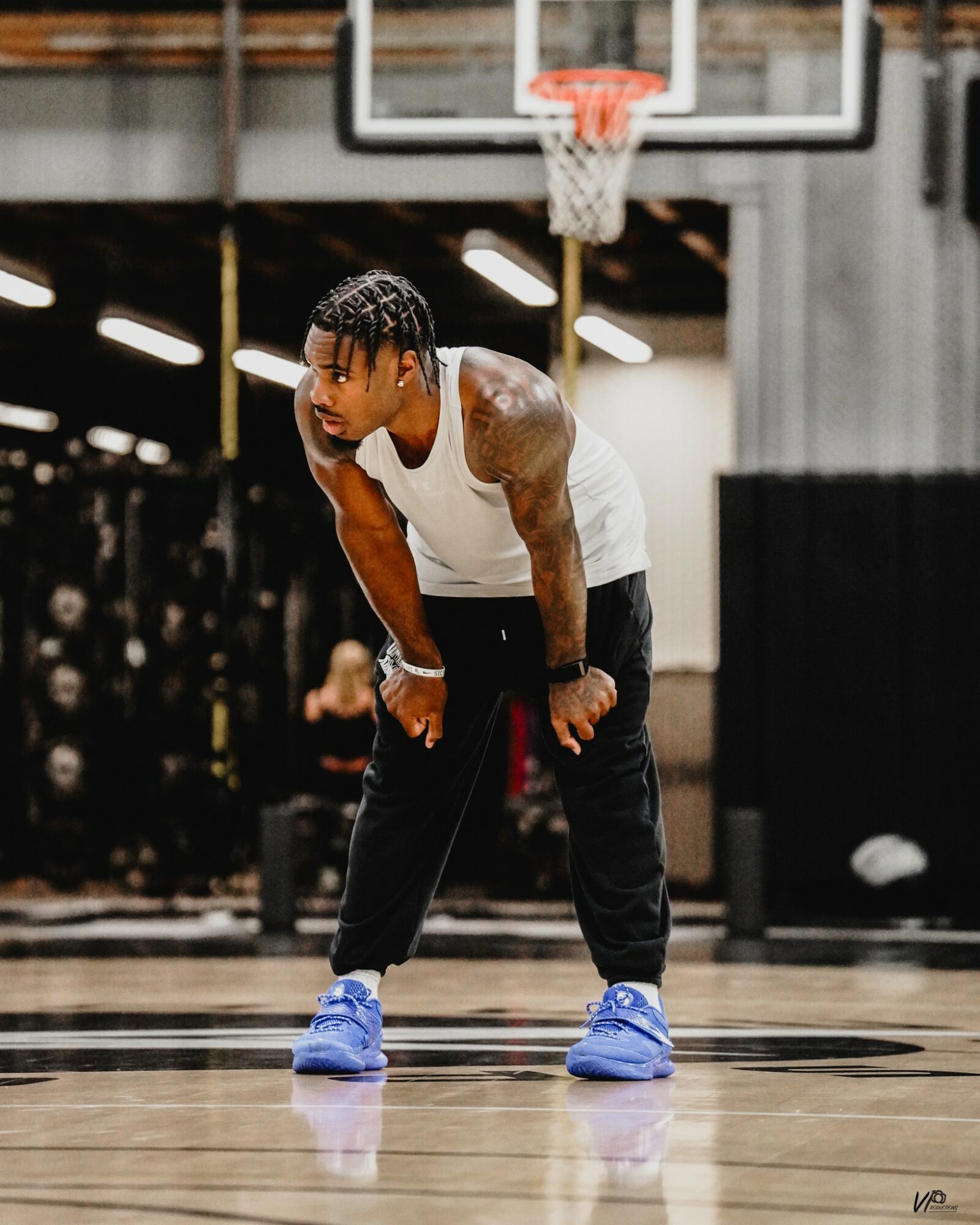 Young man in a gym, bent over with hands on knees, wearing a white tank top and blue sneakers, basketball hoop overhead.