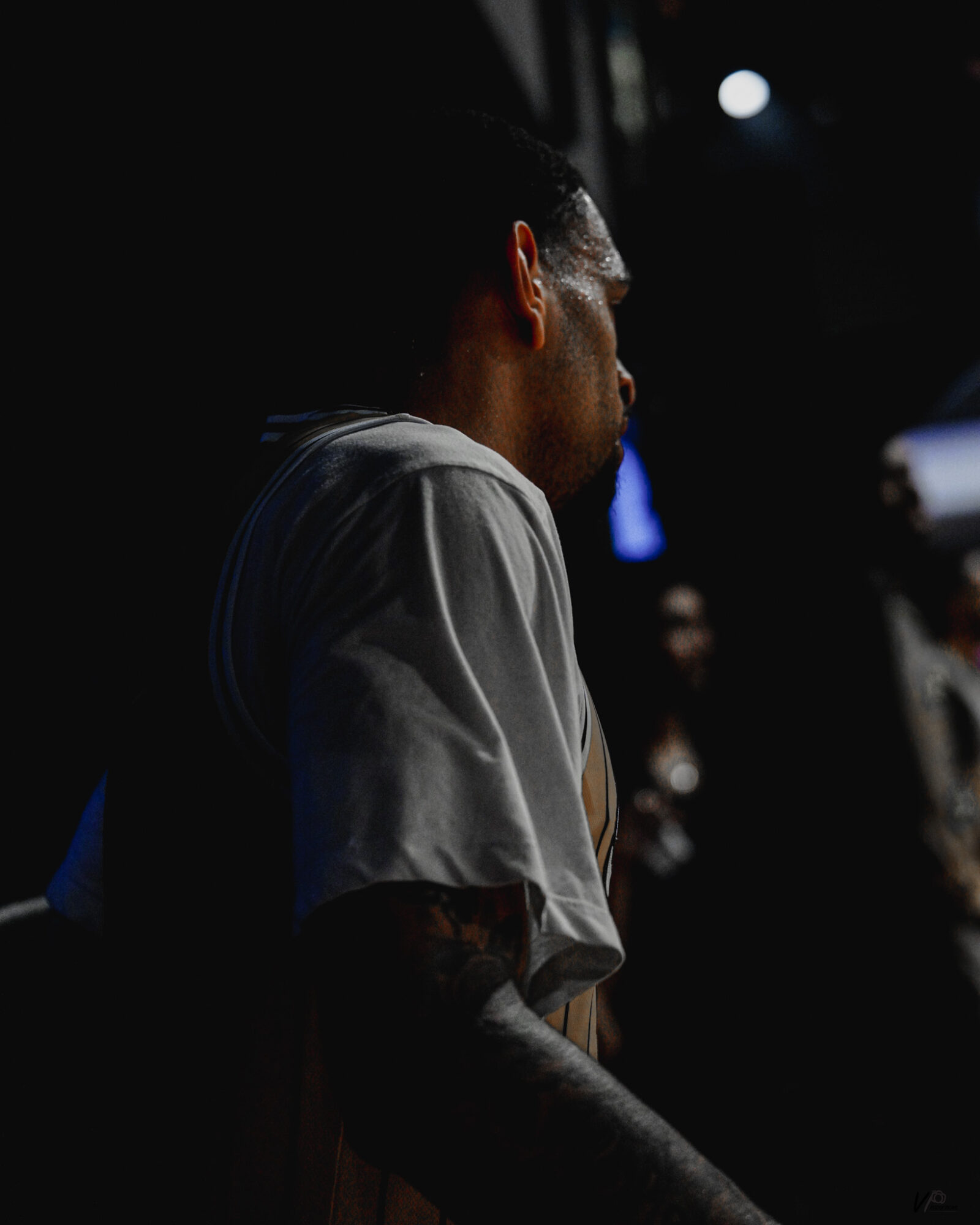 Person with short hair and tattoos on arm, wearing a light-colored shirt, in a dark environment with blurred background.