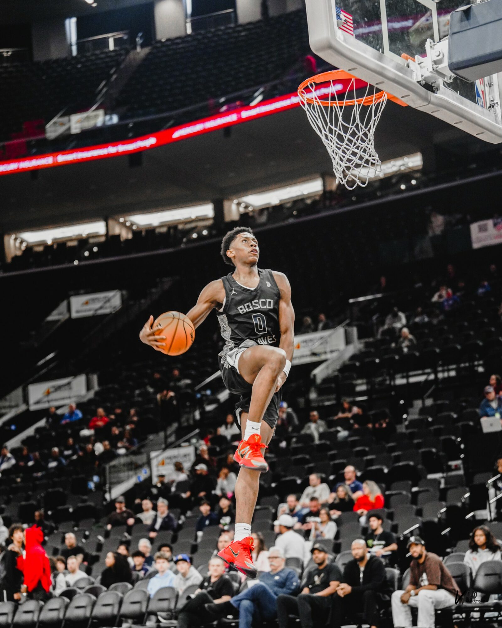 Basketball player jumping towards hoop with ball in hand during game, audience seated in background.