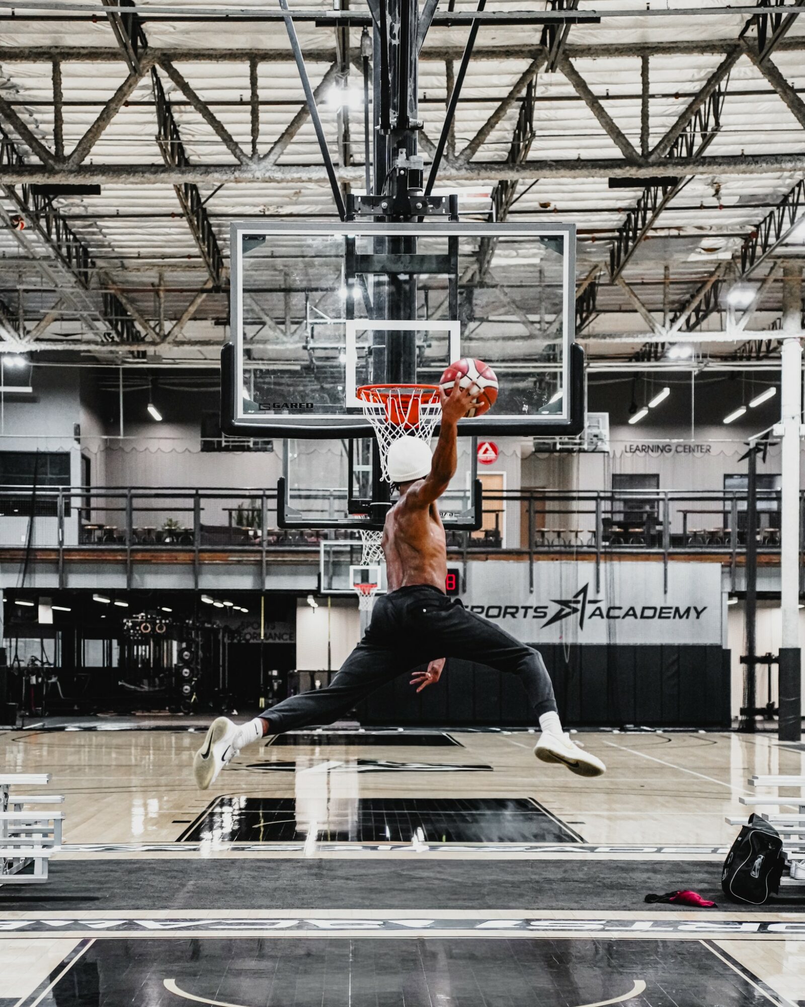 Person in a gym performing a slam dunk in front of a basketball hoop, with gym equipment and ceiling structure visible.