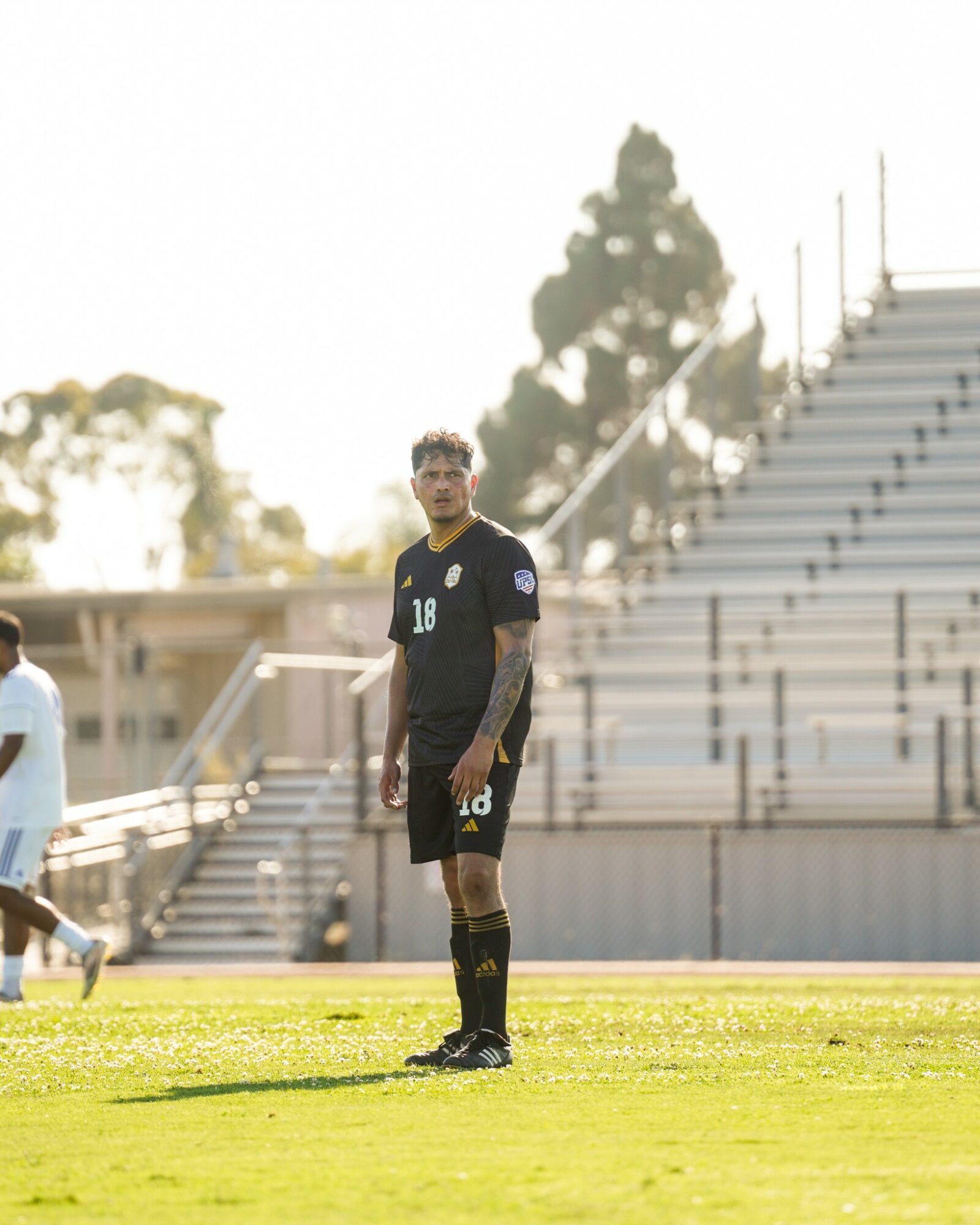 Soccer player standing on field with empty bleachers in background, sunlight behind, wearing black uniform with number 10.