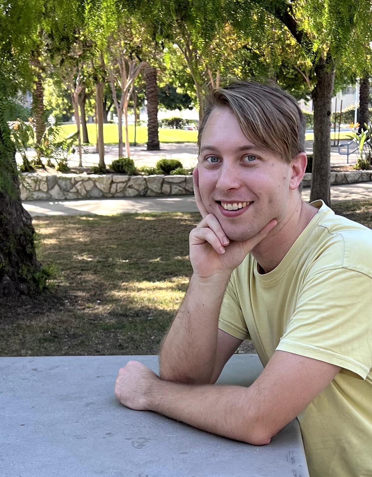 Young man with short hair smiling, resting his chin on his hand outdoors in a park, with trees and sunlight in background.