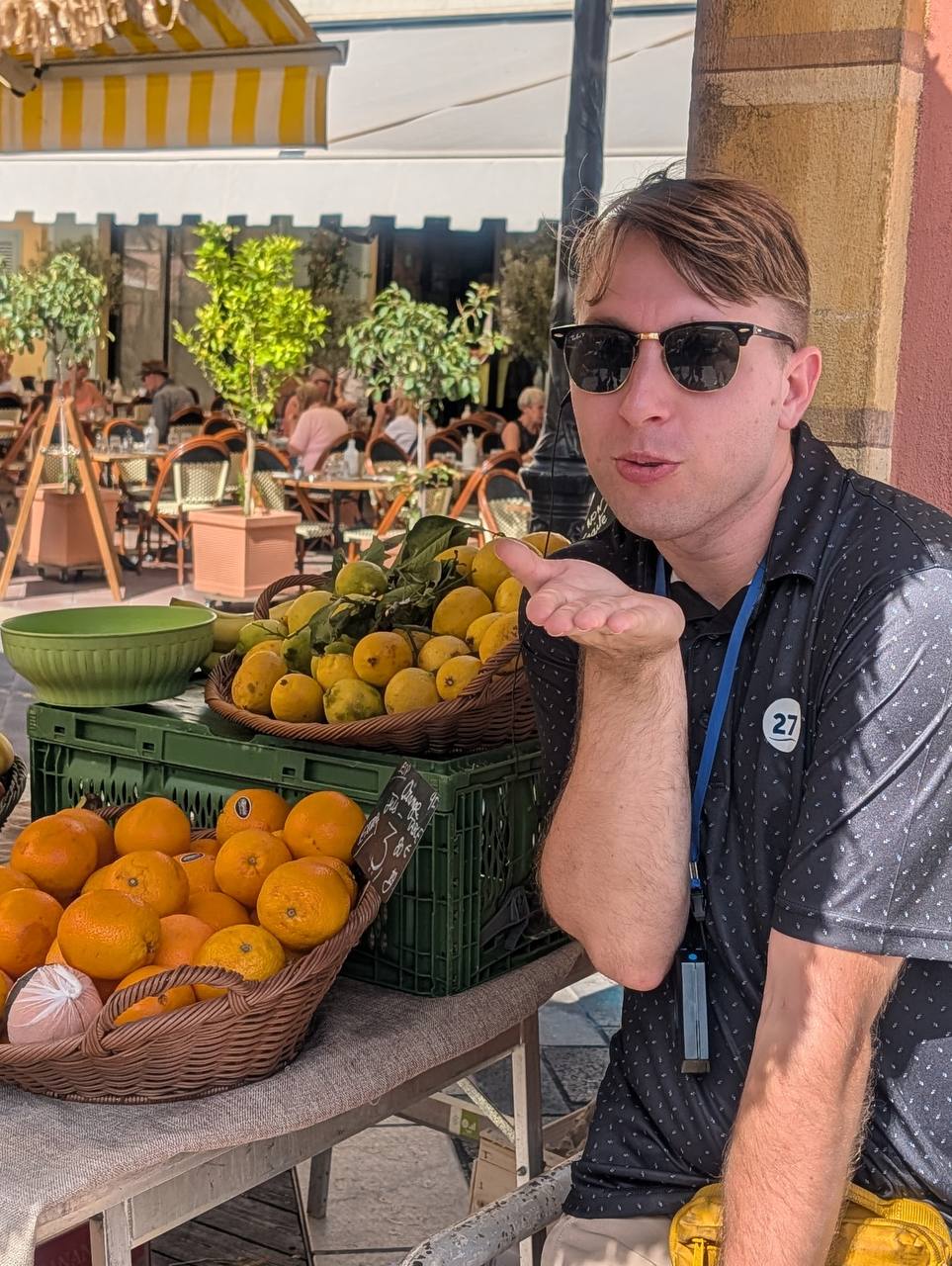 Young man wearing sunglasses and a dark shirt, sitting at an outdoor market stall with baskets of oranges and lemons.
