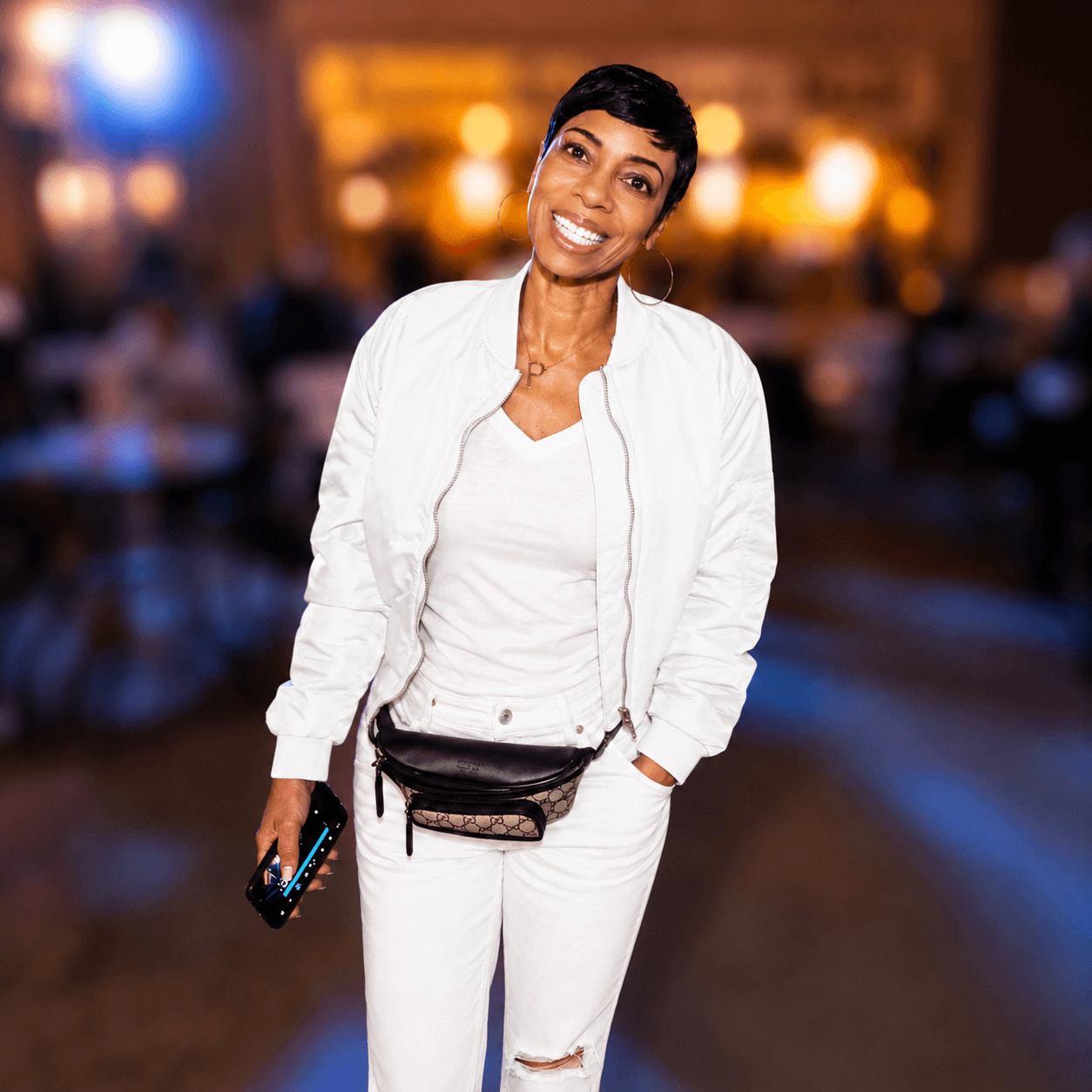 Smiling woman in white outfit holding a phone, standing in a dimly lit indoor setting with blurred background.