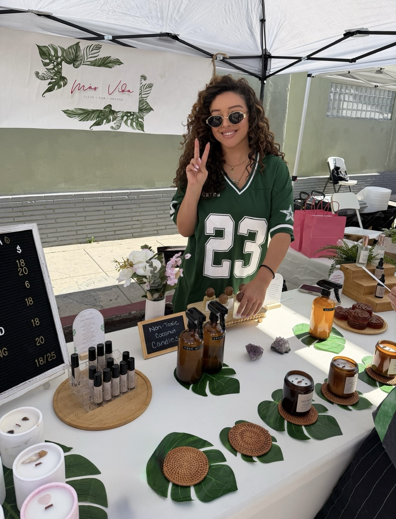 Young woman with curly hair and sunglasses smiling at a market stall, making a peace sign, wearing a green jersey with number 23.