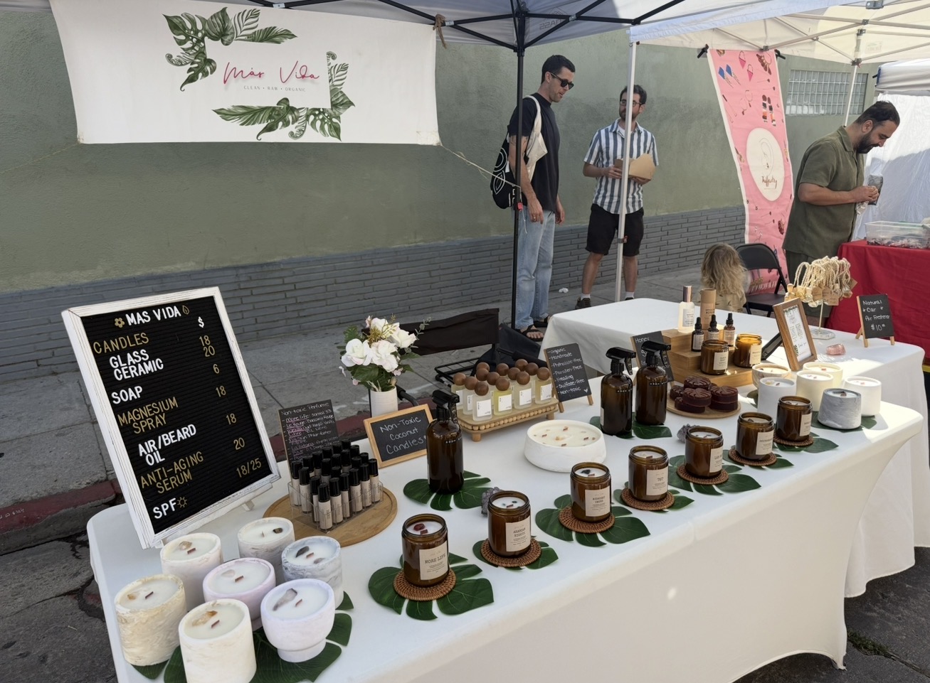 Table with jars, candles, and a blackboard menu at an outdoor market stall, with three people browsing in the background.