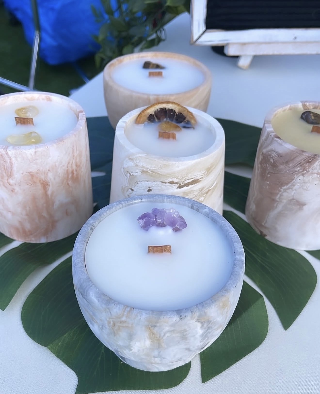 Five candles with different colors and decorations on large green leaves, outdoors setting, with a blurred background.