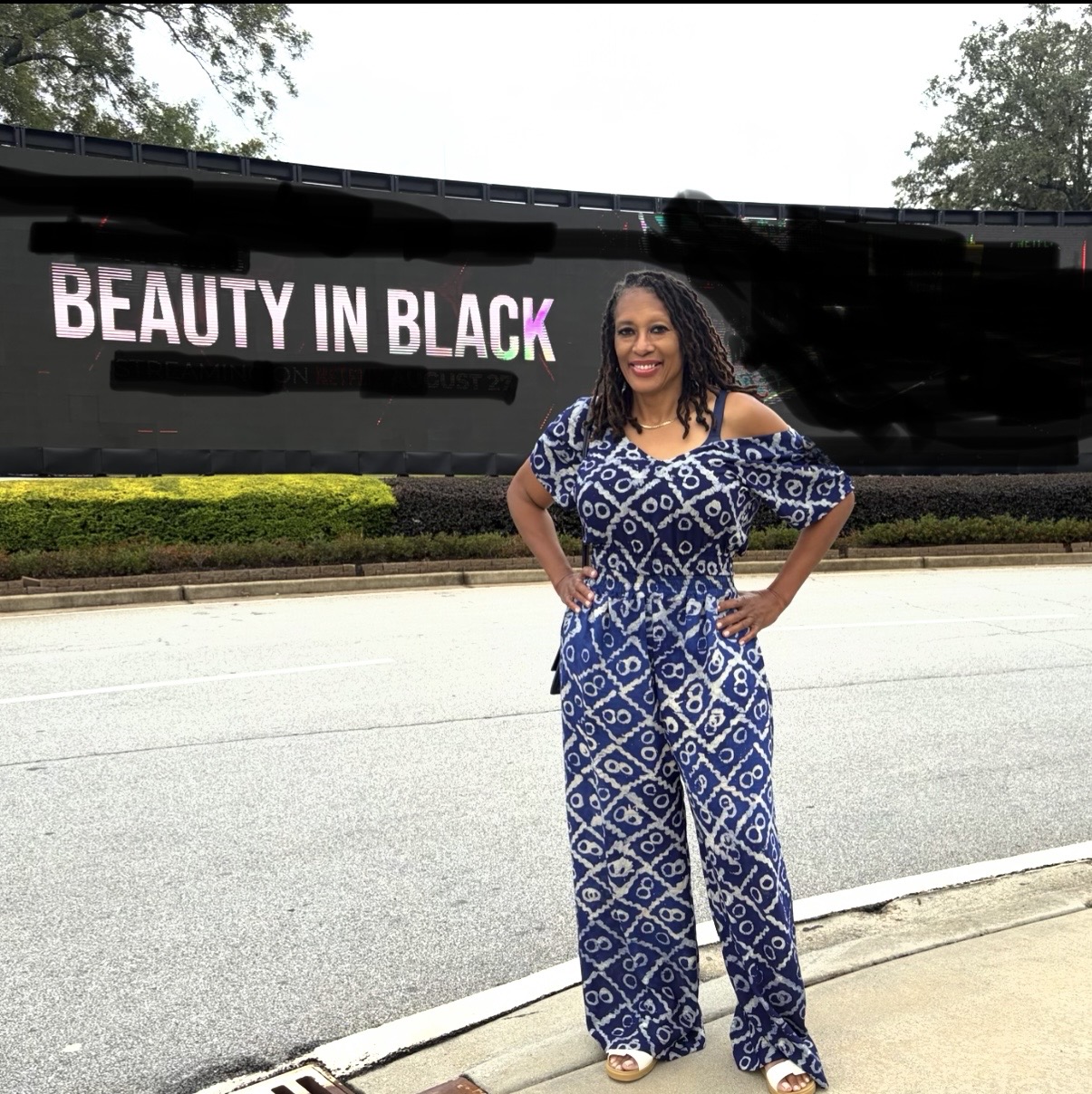 Woman in blue patterned jumpsuit standing on sidewalk in front of a black wall with pink and white text, smiling.