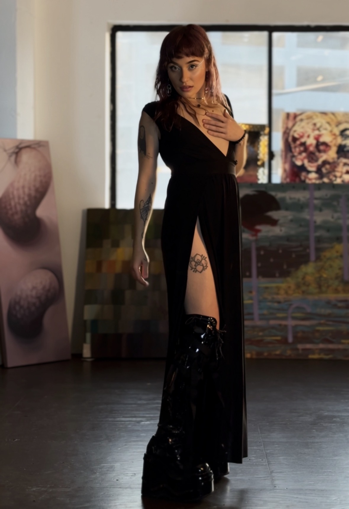 Woman with red hair in a black dress with a high slit, standing indoors near large windows, holding her chest. Word count: 20
