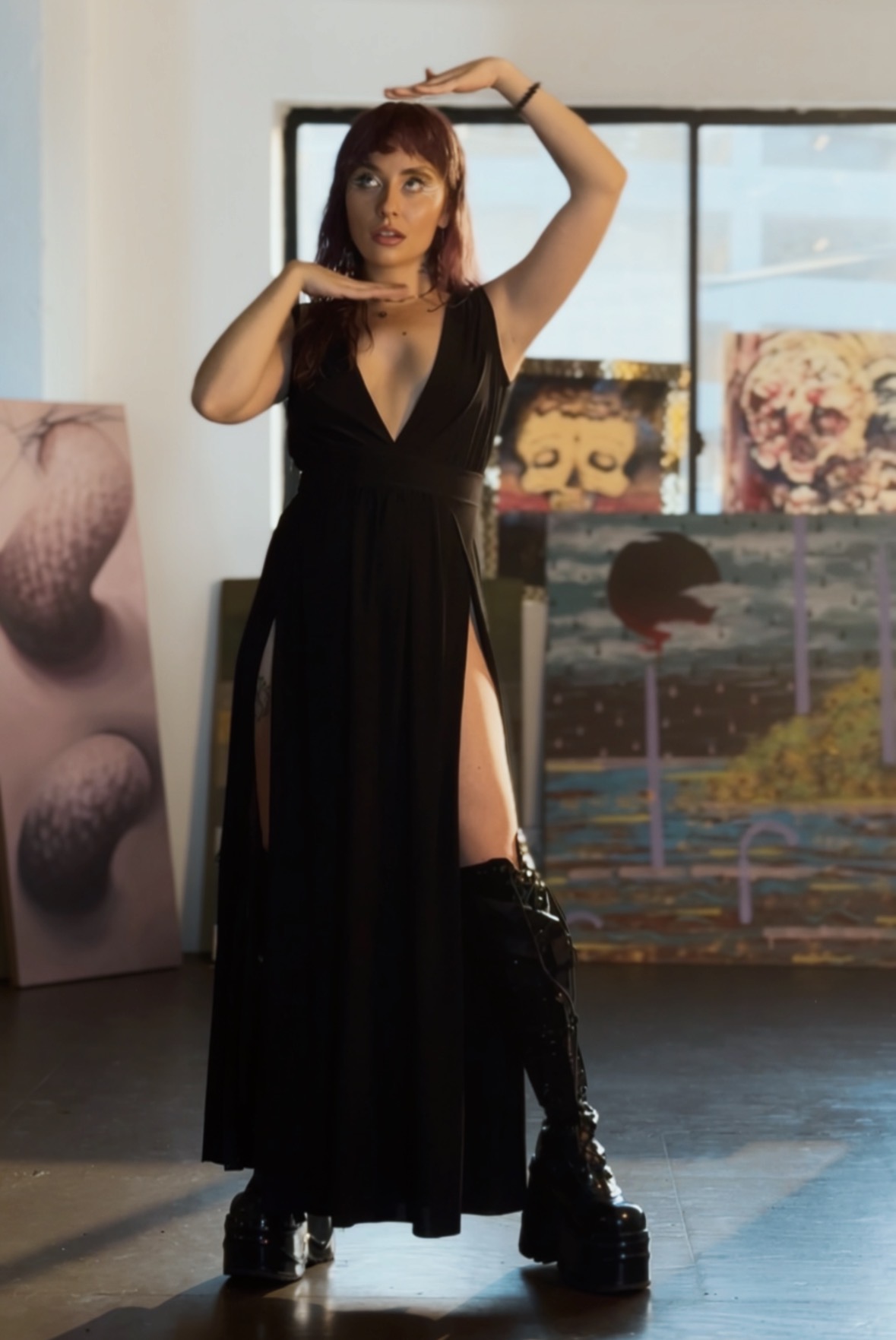 Woman in black dress with high slit, standing indoors with artwork in background, one arm raised above head, other near face.