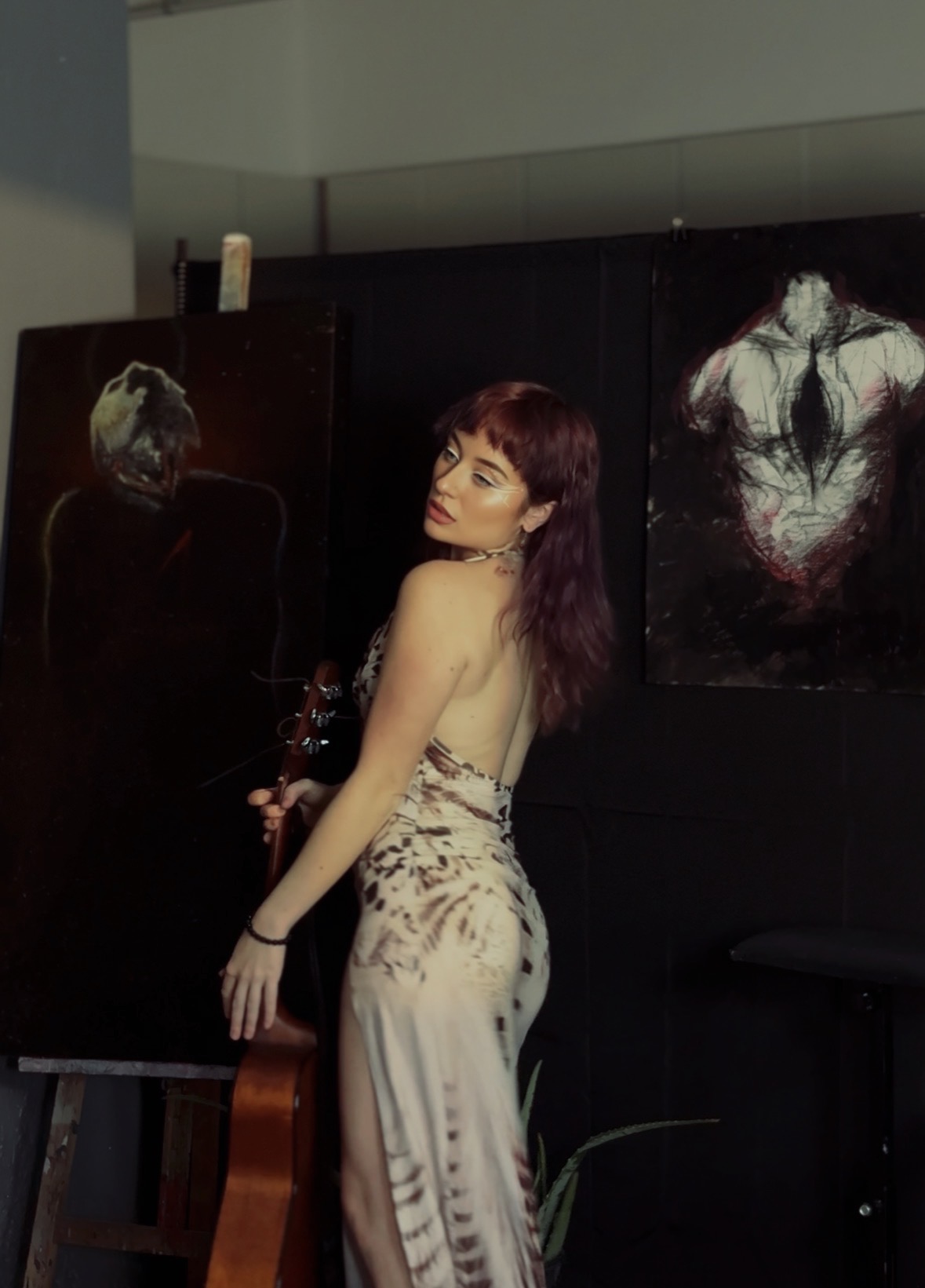 Young woman with purple hair stands in front of dark paintings, looking over her shoulder, in an art gallery setting.