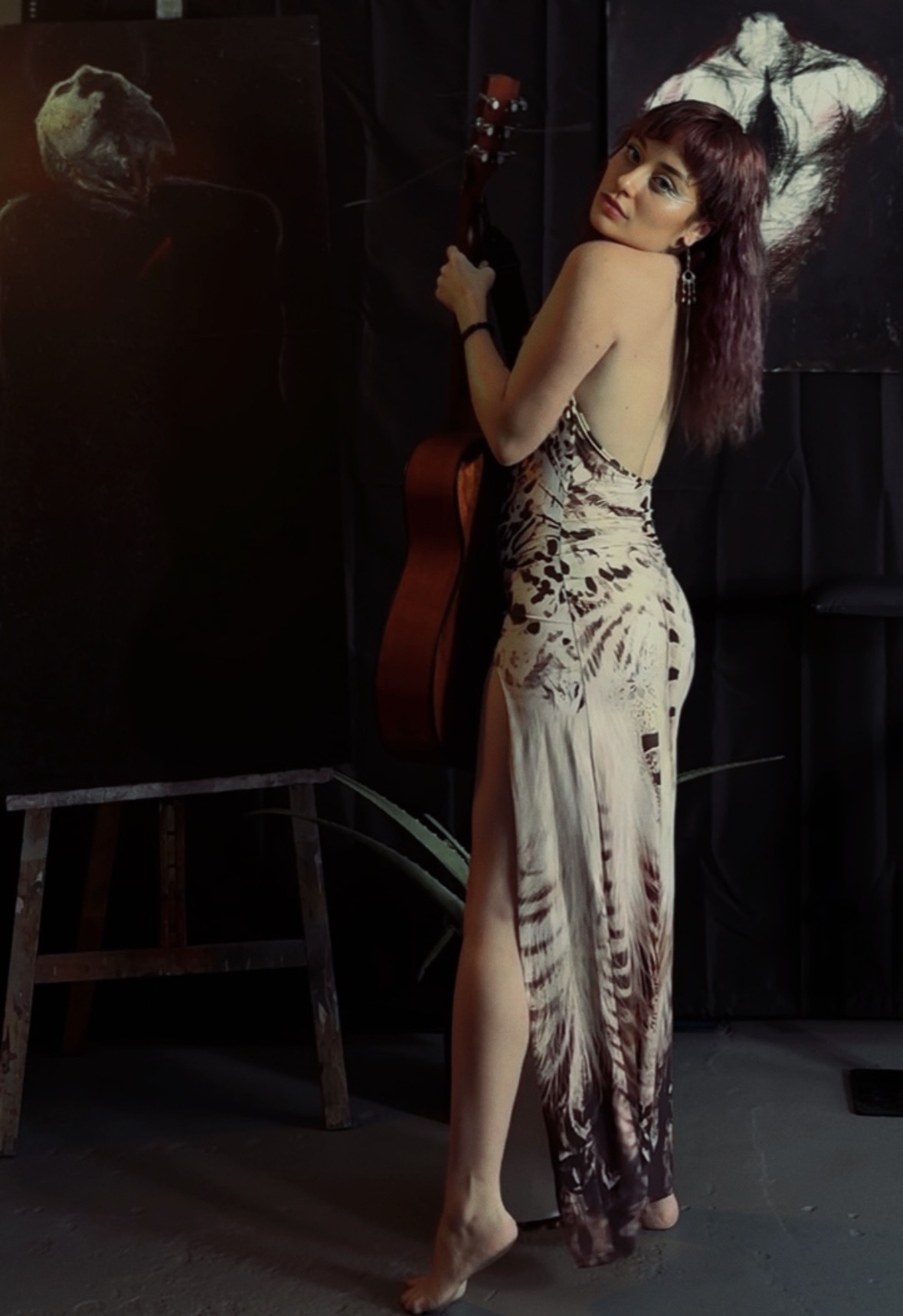 Woman in a patterned dress holding a guitar, standing in a dark room with artwork on the wall.
