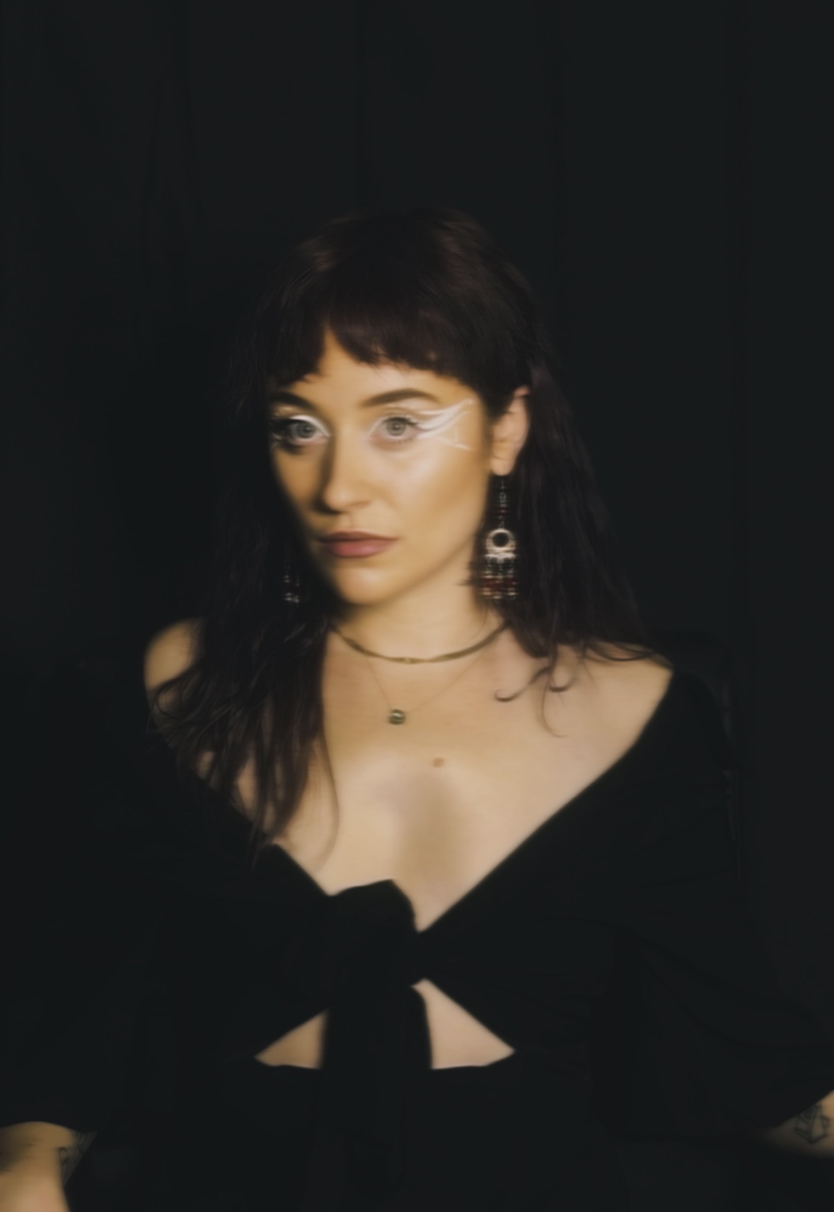 Woman with dark hair and earrings, wearing a black top with a bow, against a dark background.