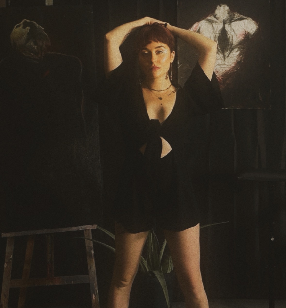 Young woman with dark hair and makeup, wearing a black outfit, standing with arms raised behind her head in a dimly lit room.