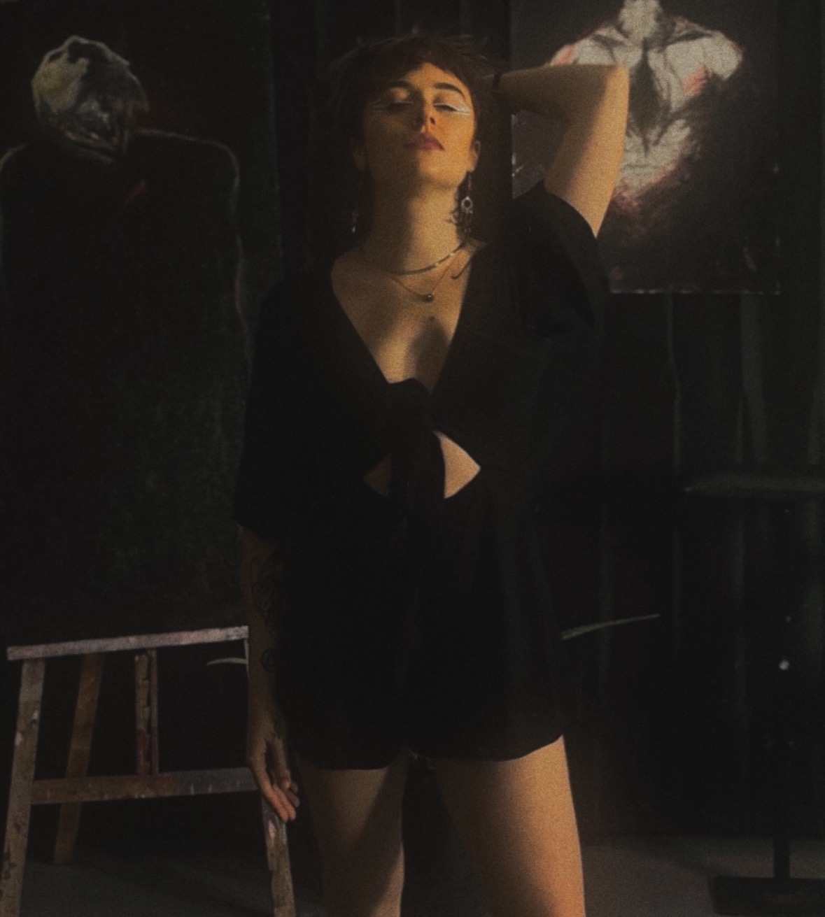 A woman with dark hair and makeup poses with one arm raised, wearing a black outfit in a dimly lit room.