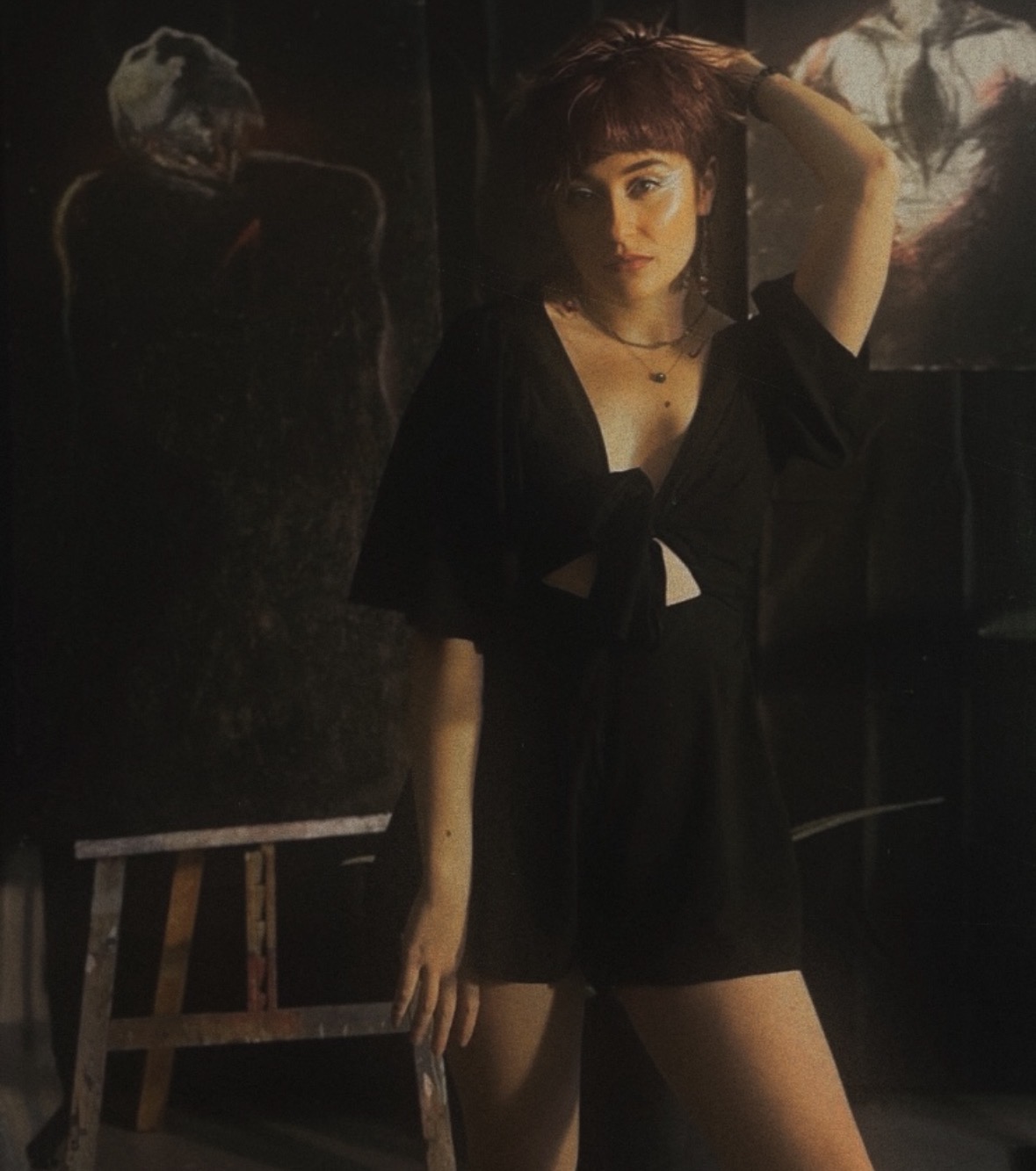 Young woman with short hair and dark clothing, standing indoors with a dark background and artwork behind her.