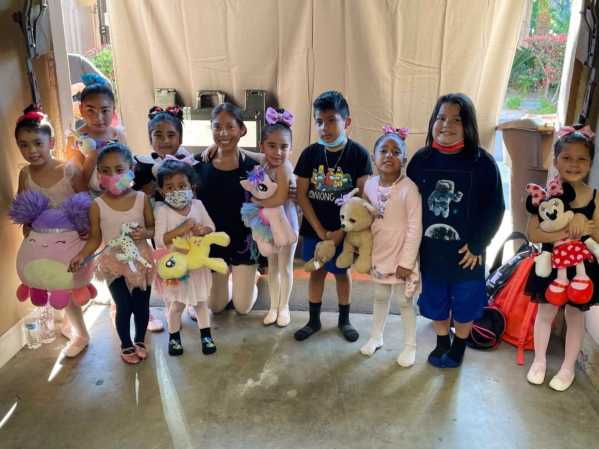 Group of children holding plush toys, standing indoors in front of curtains, some wearing masks and colorful outfits.