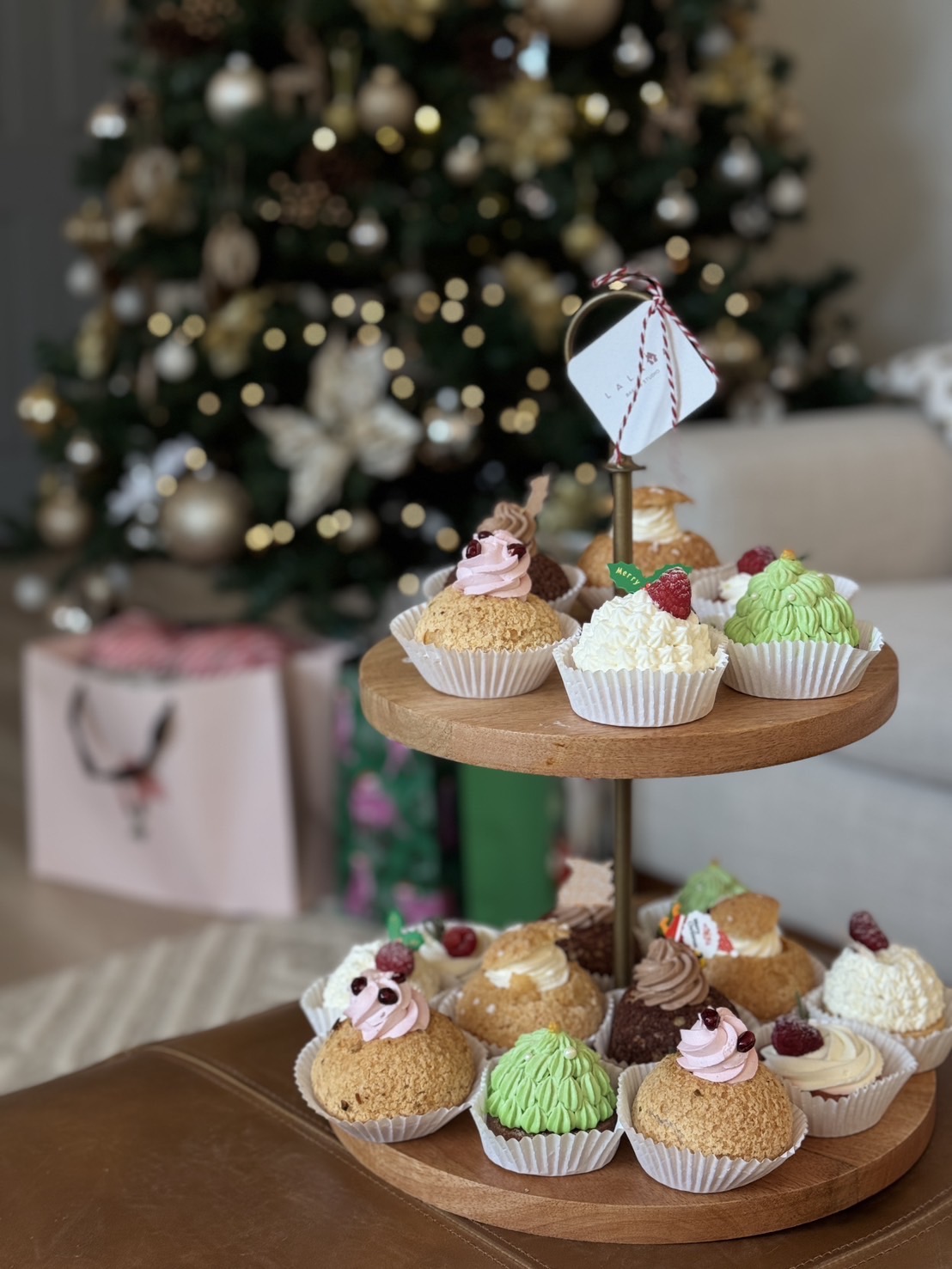 Cupcakes with colorful frosting and decorations on a two-tiered wooden stand in front of a decorated Christmas tree.
