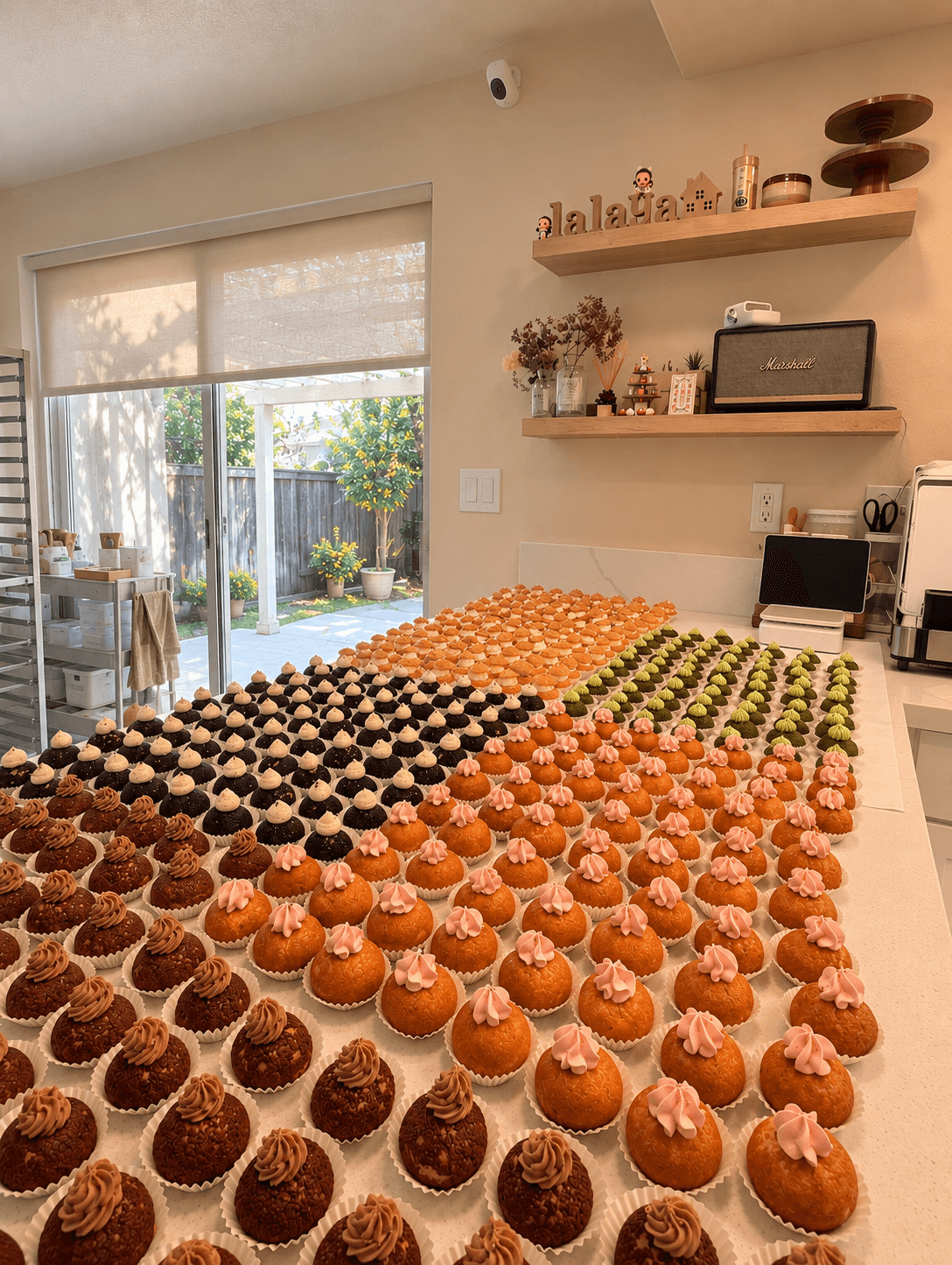 Table filled with colorful cupcakes in a bright kitchen, with a sliding glass door leading to a garden outside.
