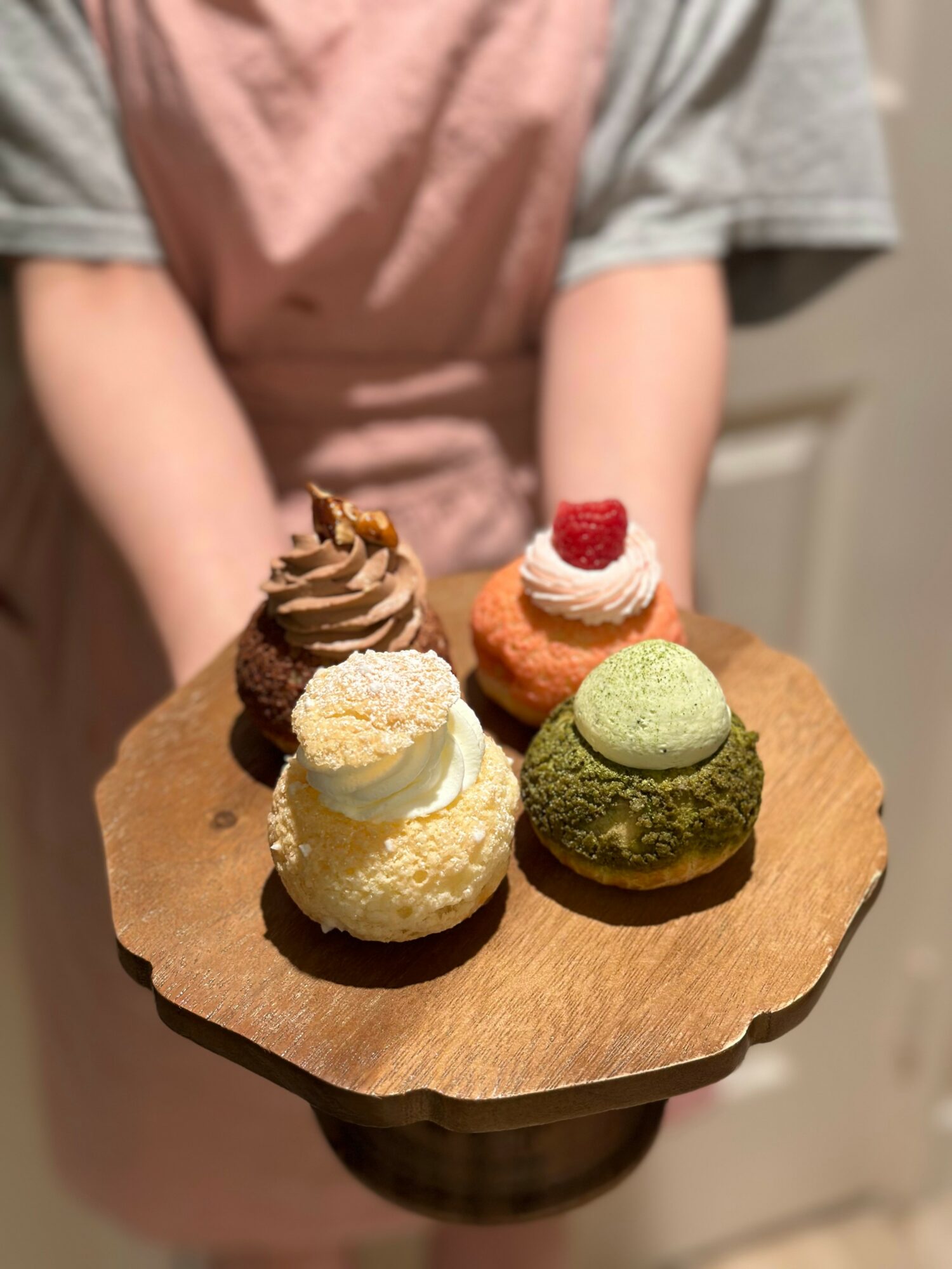 Person holding a wooden platter with five decorated cupcakes, wearing a pink apron and gray shirt, in a kitchen.