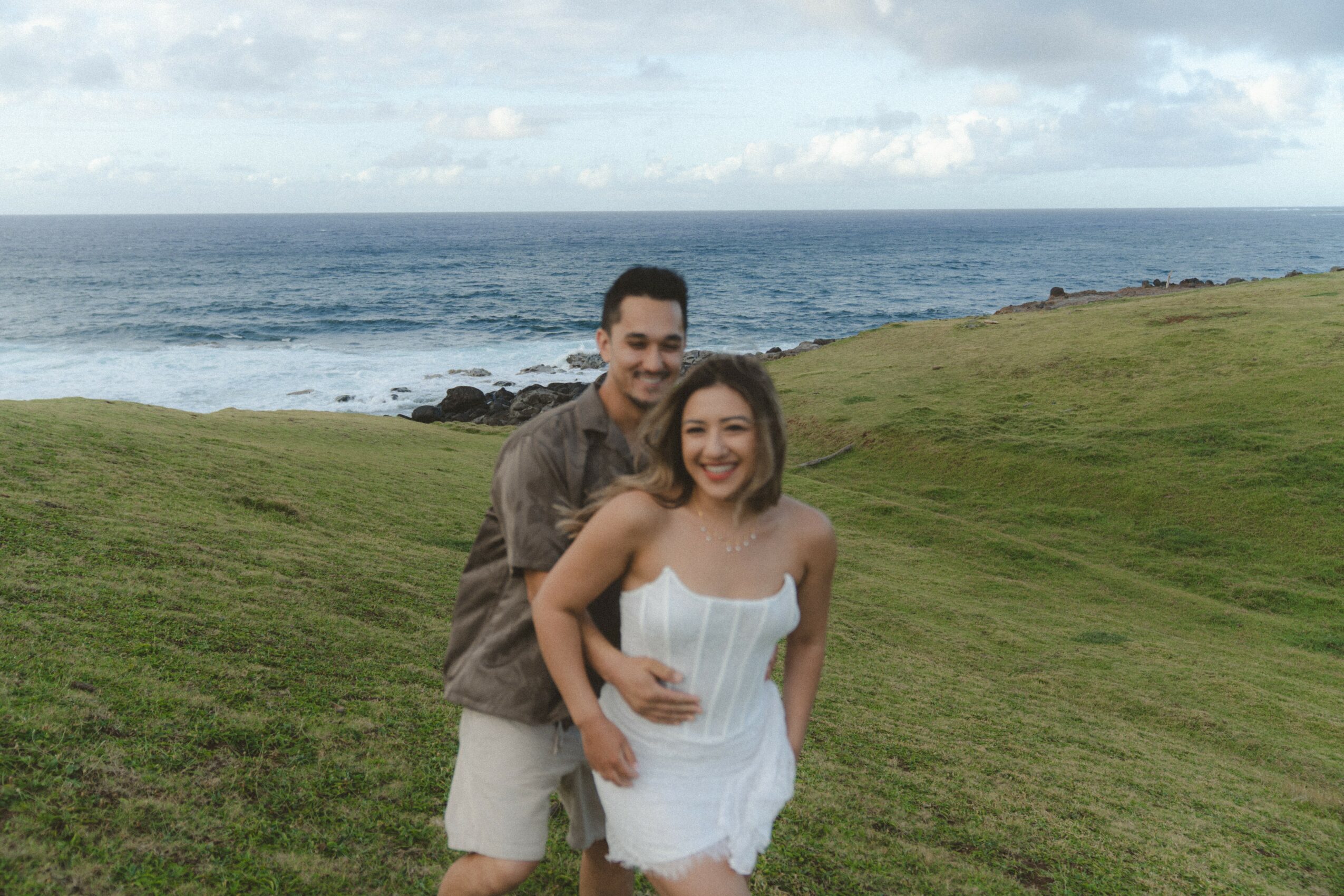 A smiling couple stands on a grassy area near the ocean, with waves and a cloudy sky in the background.
