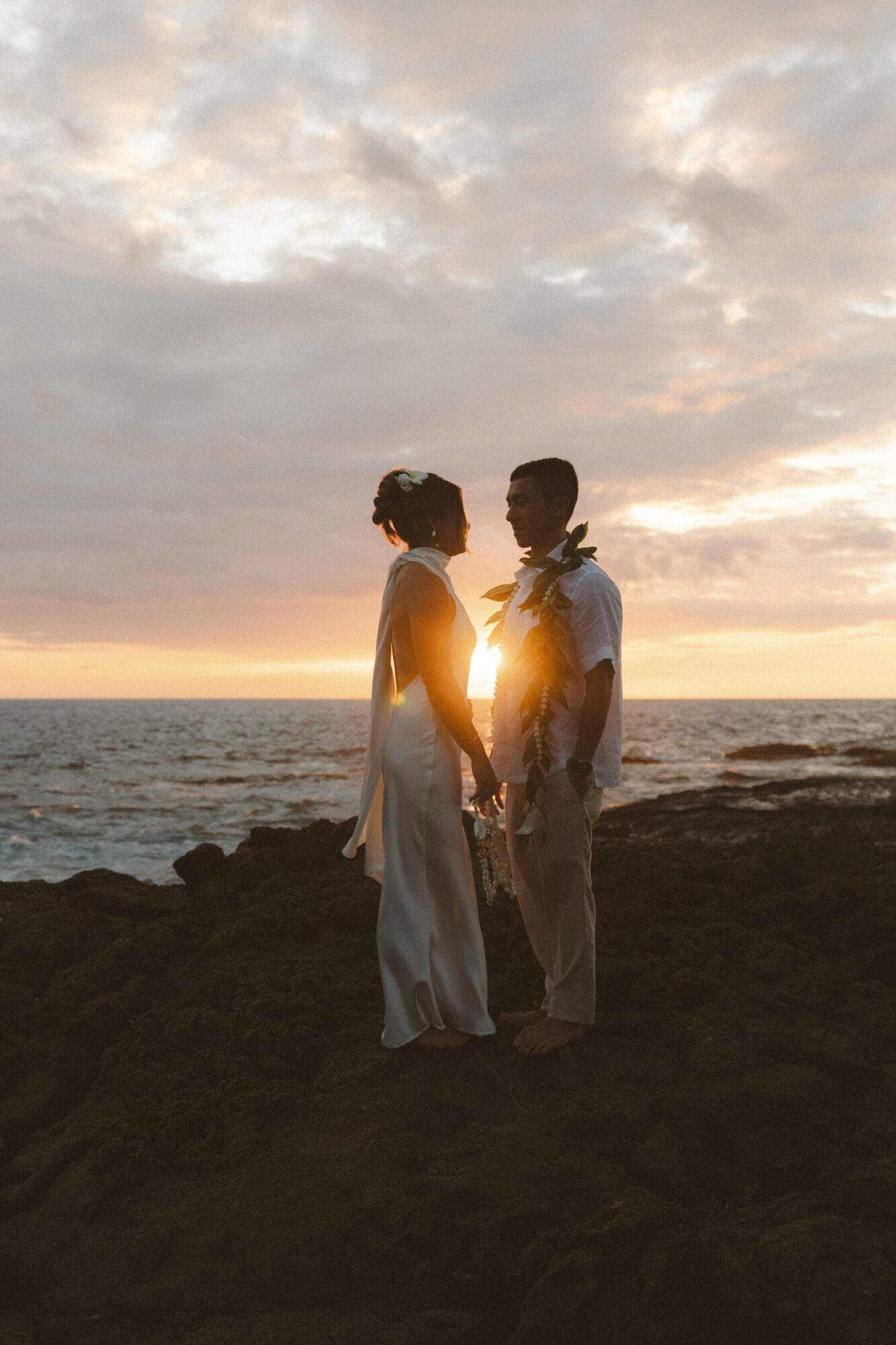 Two people stand on a rocky beach at sunset, facing each other with the sun setting behind them.