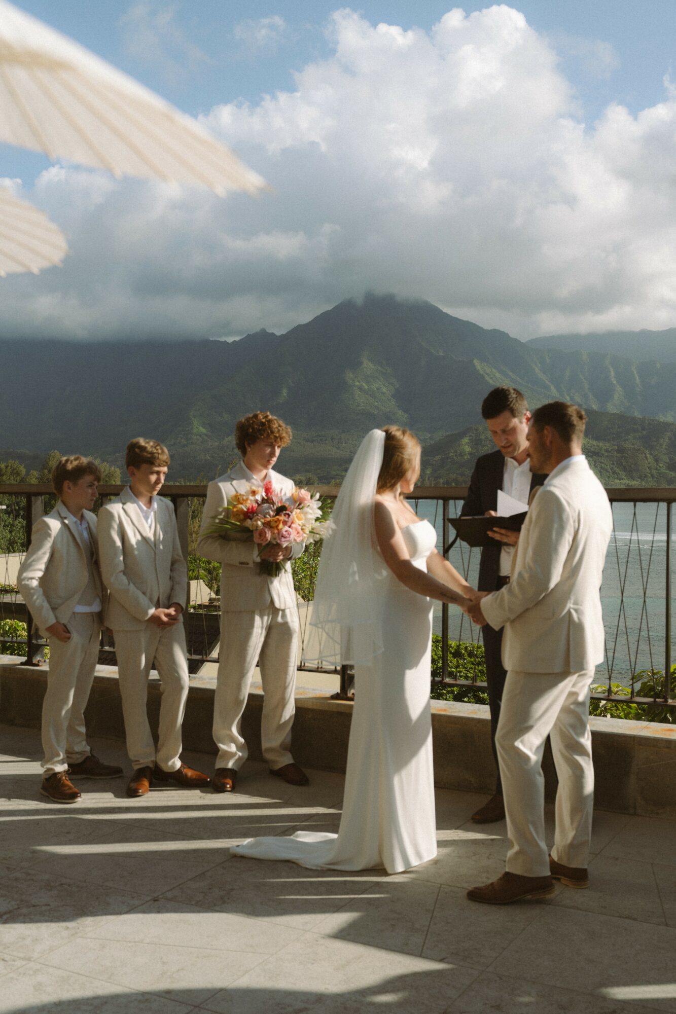 A wedding ceremony outdoors with a mountain view, bride and groom holding hands, officiant reading, group of children in formal attire, cloudy sky.