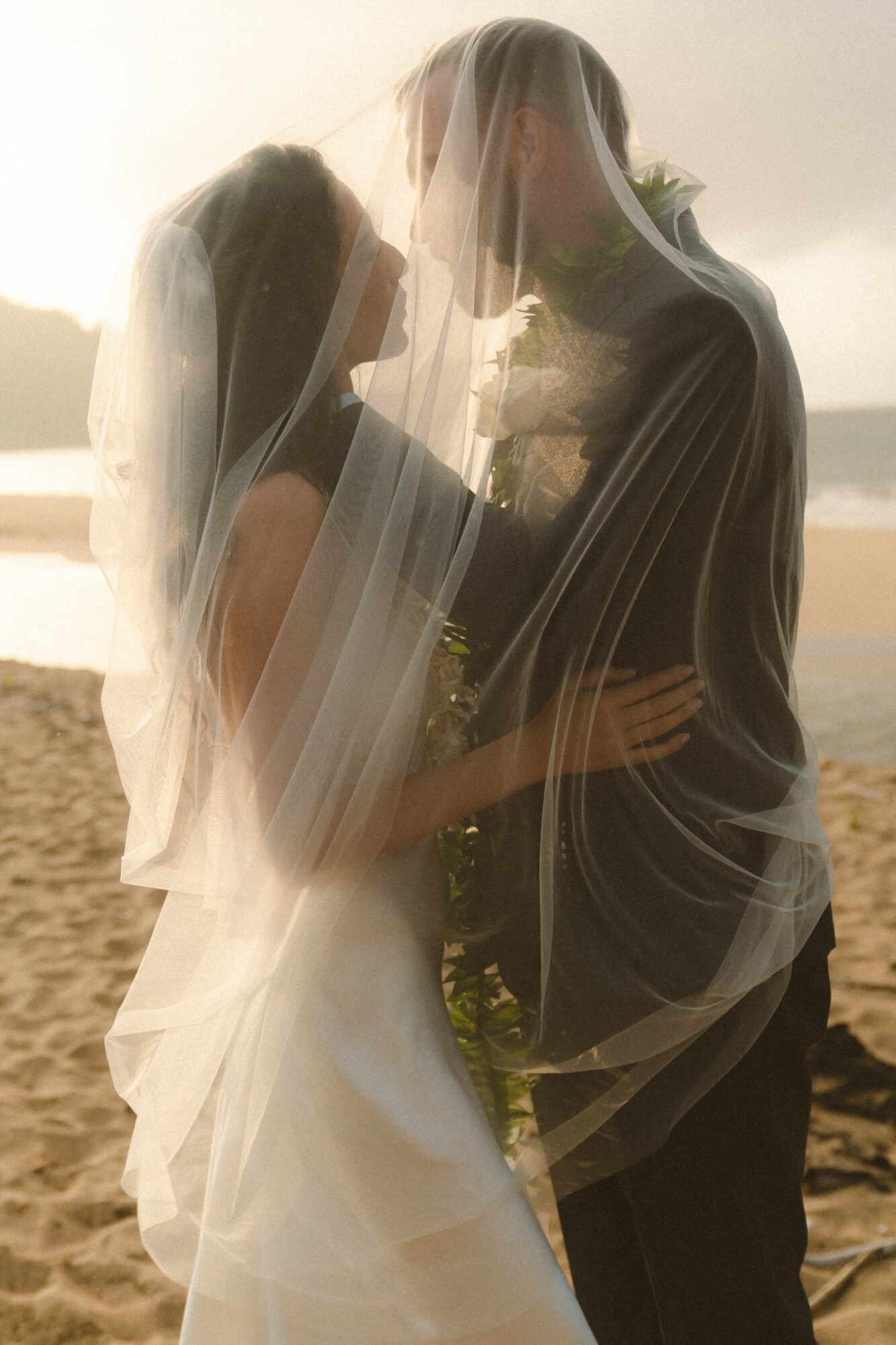 A bride and groom stand close together on a beach, with a veil covering both of them, backlit by sunlight.