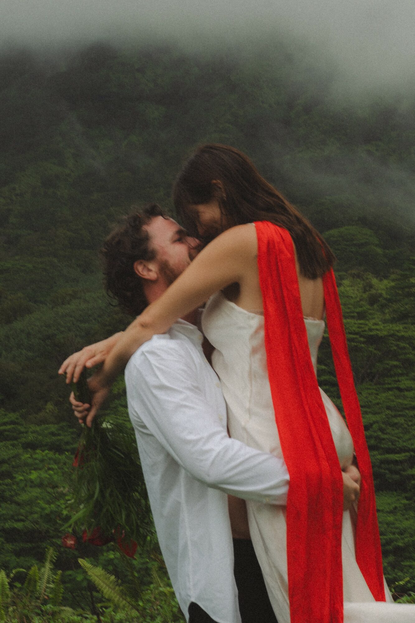Two people embrace outdoors with a green, foggy landscape in the background, one wearing a white outfit and the other a white dress with a red scarf.