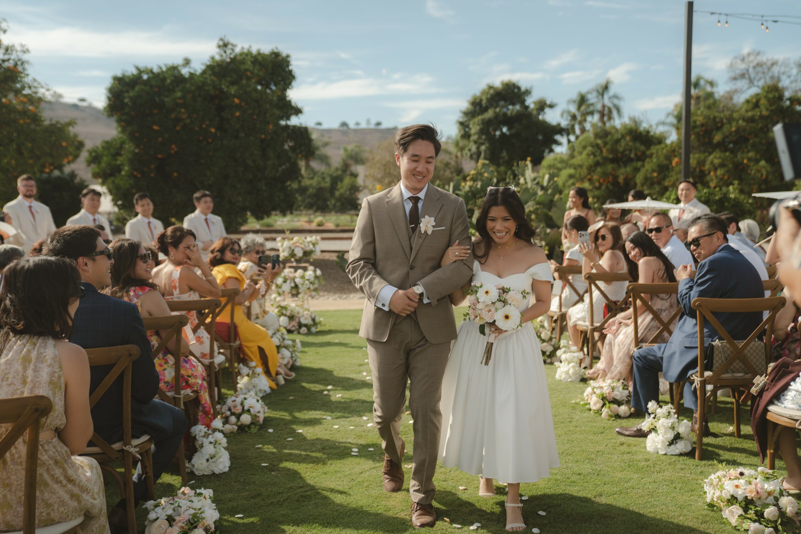 A wedding ceremony outdoors with a bride and groom walking down the aisle, guests seated on both sides, trees and sky in background.